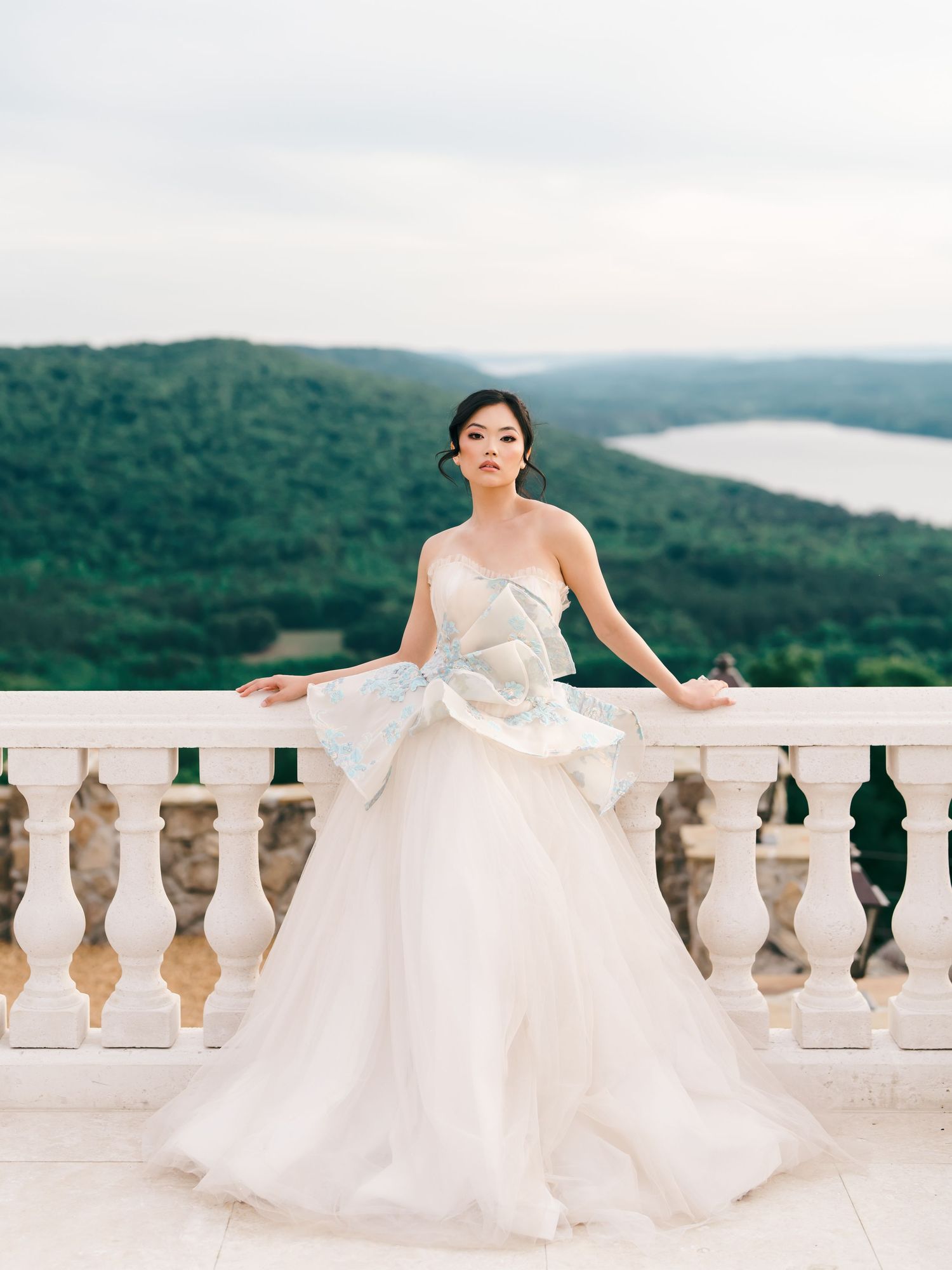 Bride in strapless white ballgown with blue bow on scenic balcony overlooking lush green hills and lake.