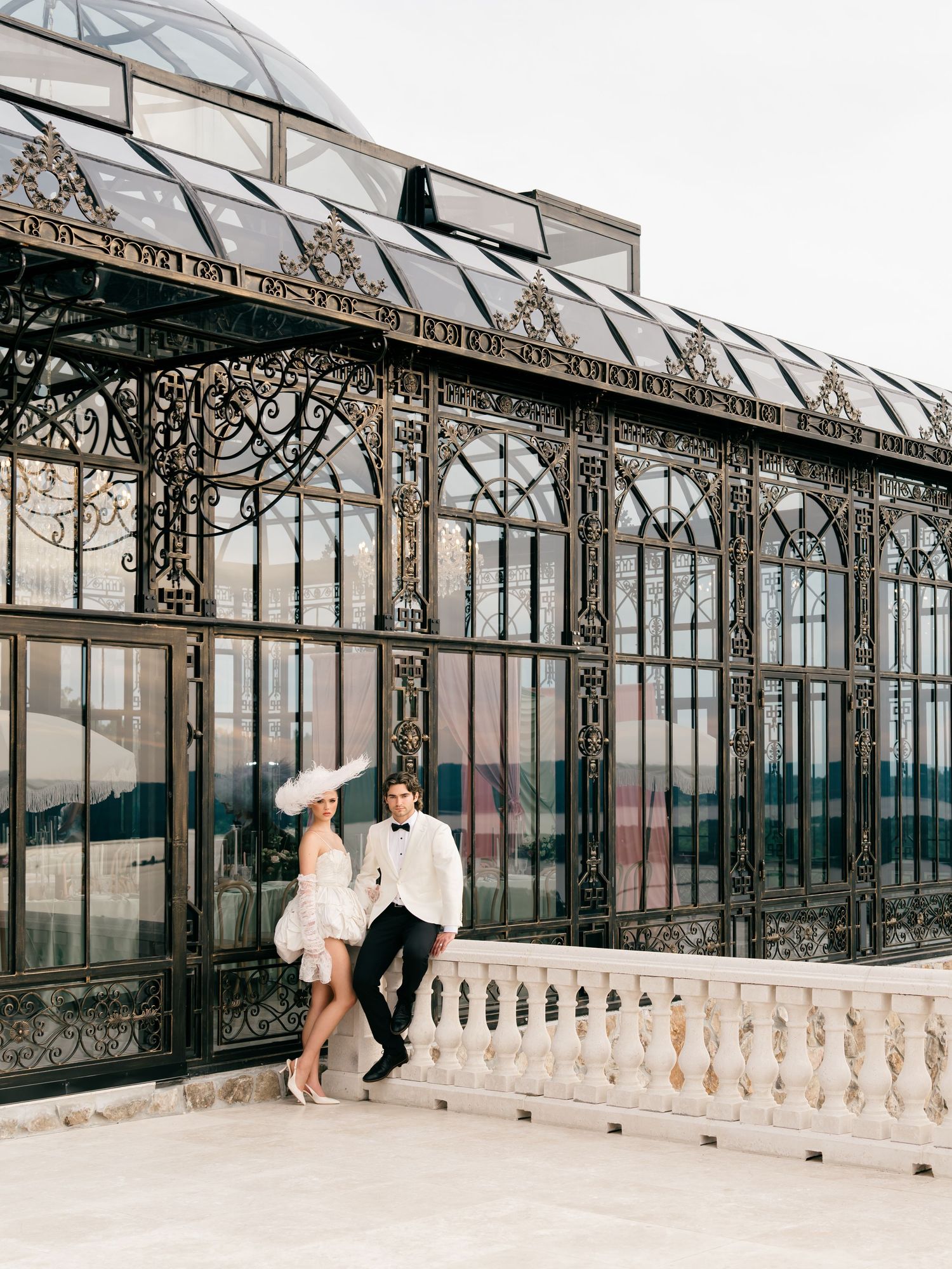 Elegant couple in formal attire posing before ornate Victorian greenhouse with intricate ironwork and glass dome.