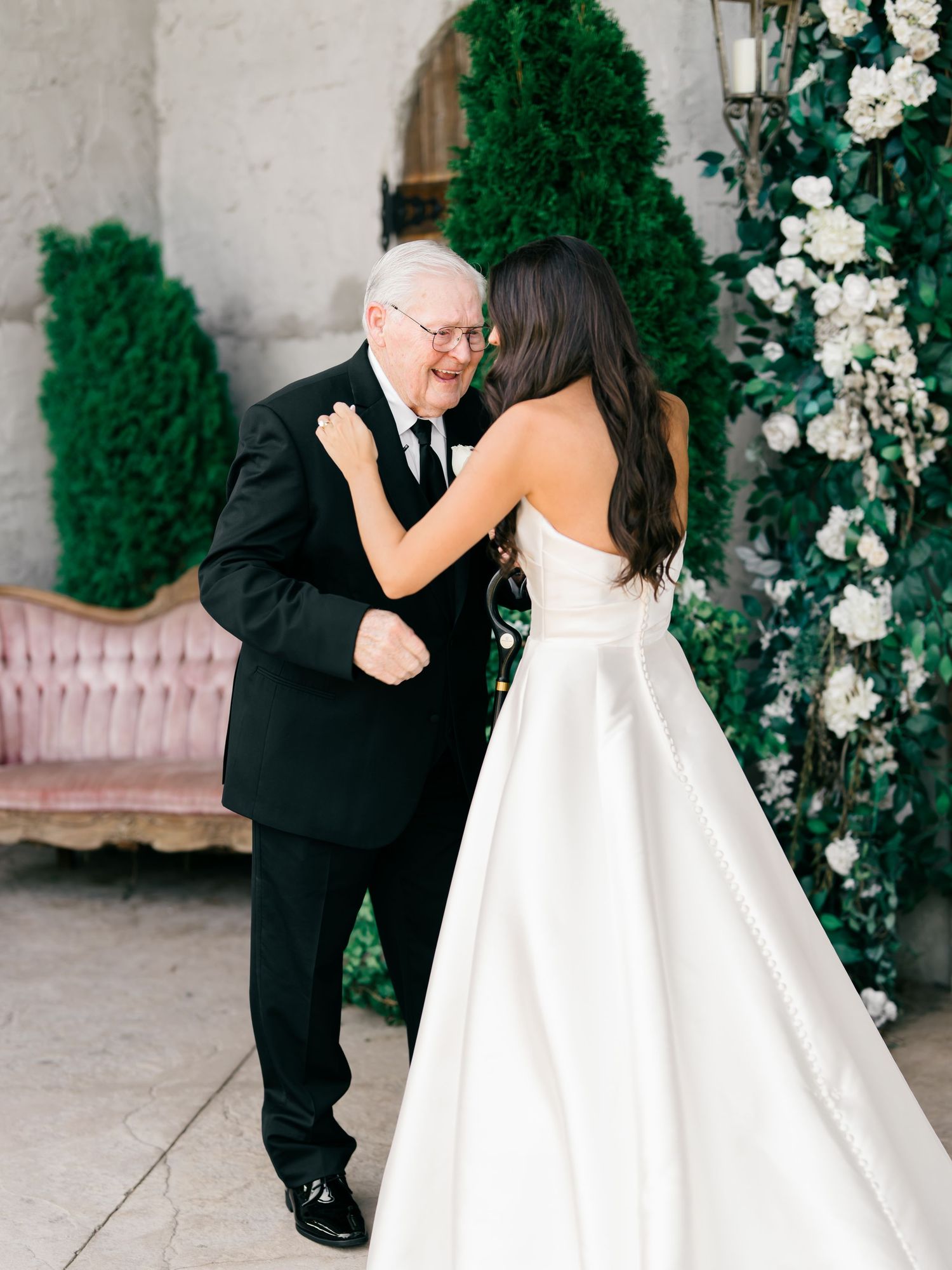 A couple in formal wedding attire dances and twirls together in a garden setting with white floral decorations.