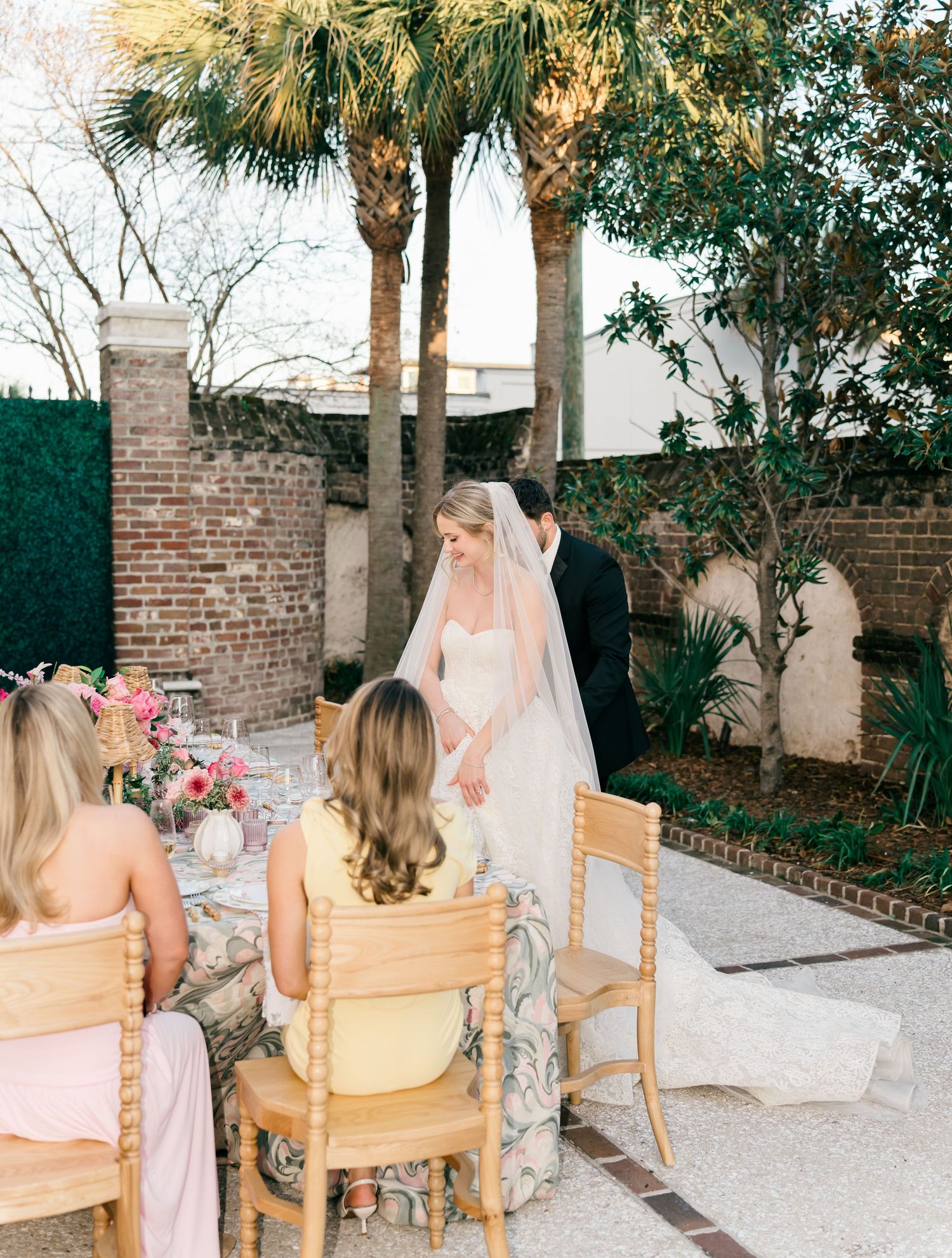 Wedding party gathered in an outdoor garden setting with palm trees and brick walls, seated on wooden chairs.