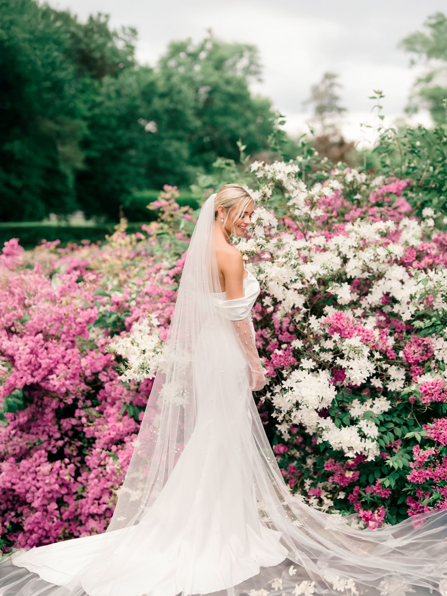A bride in a flowing white wedding dress and veil stands in a garden filled with blooming pink and white flowers.