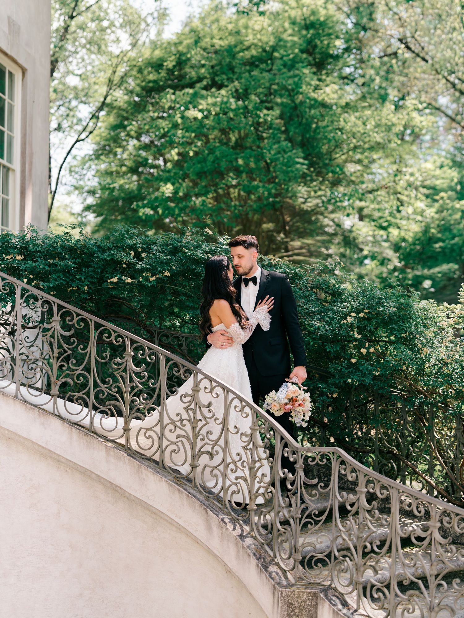 Bride and groom share a romantic kiss on an ornate wrought iron staircase surrounded by lush green trees.