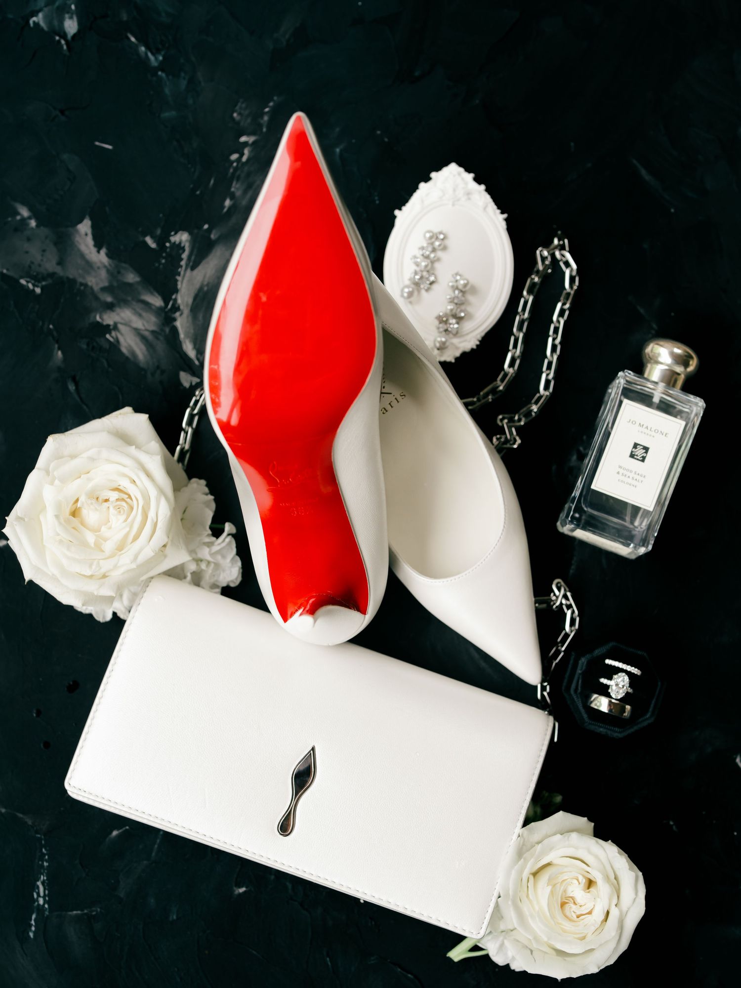 White Christian Louboutin heels with red soles, white clutch, roses, jewelry, and Jo Malone perfume on dark marble.
