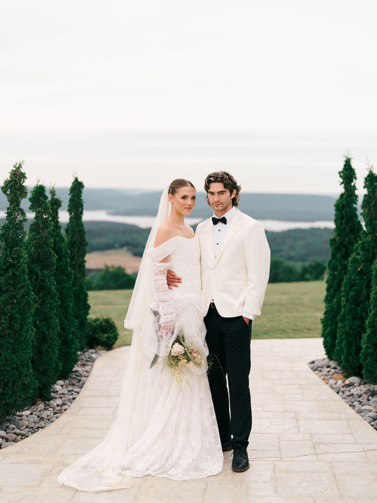 Bride in white gown with veil and groom in white tuxedo jacket pose on stone path with evergreen trees and lake view.