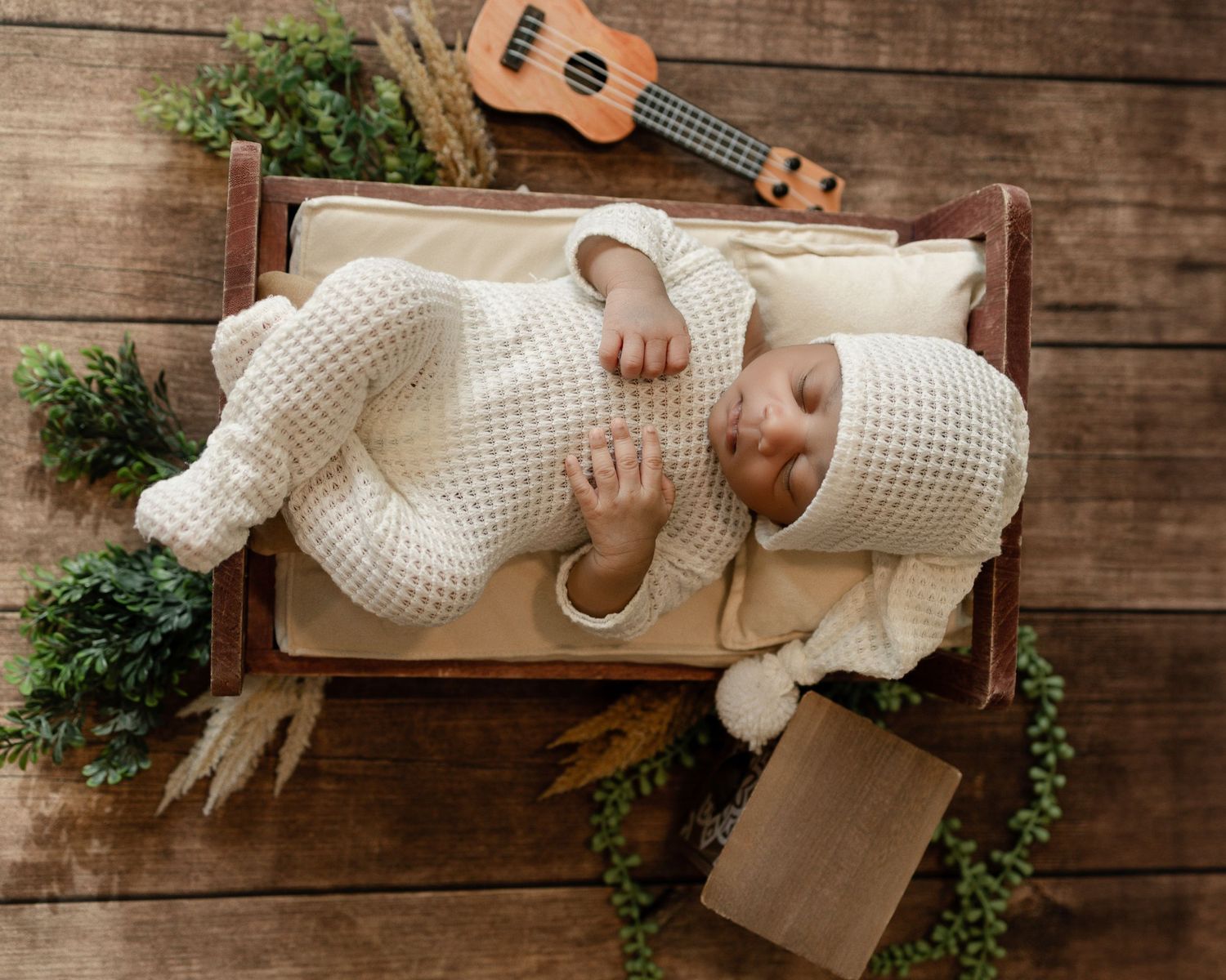 A cozy rustic newborn photo setup featuring a small bed with white bedding, ukulele, and greenery on wooden backdrop.