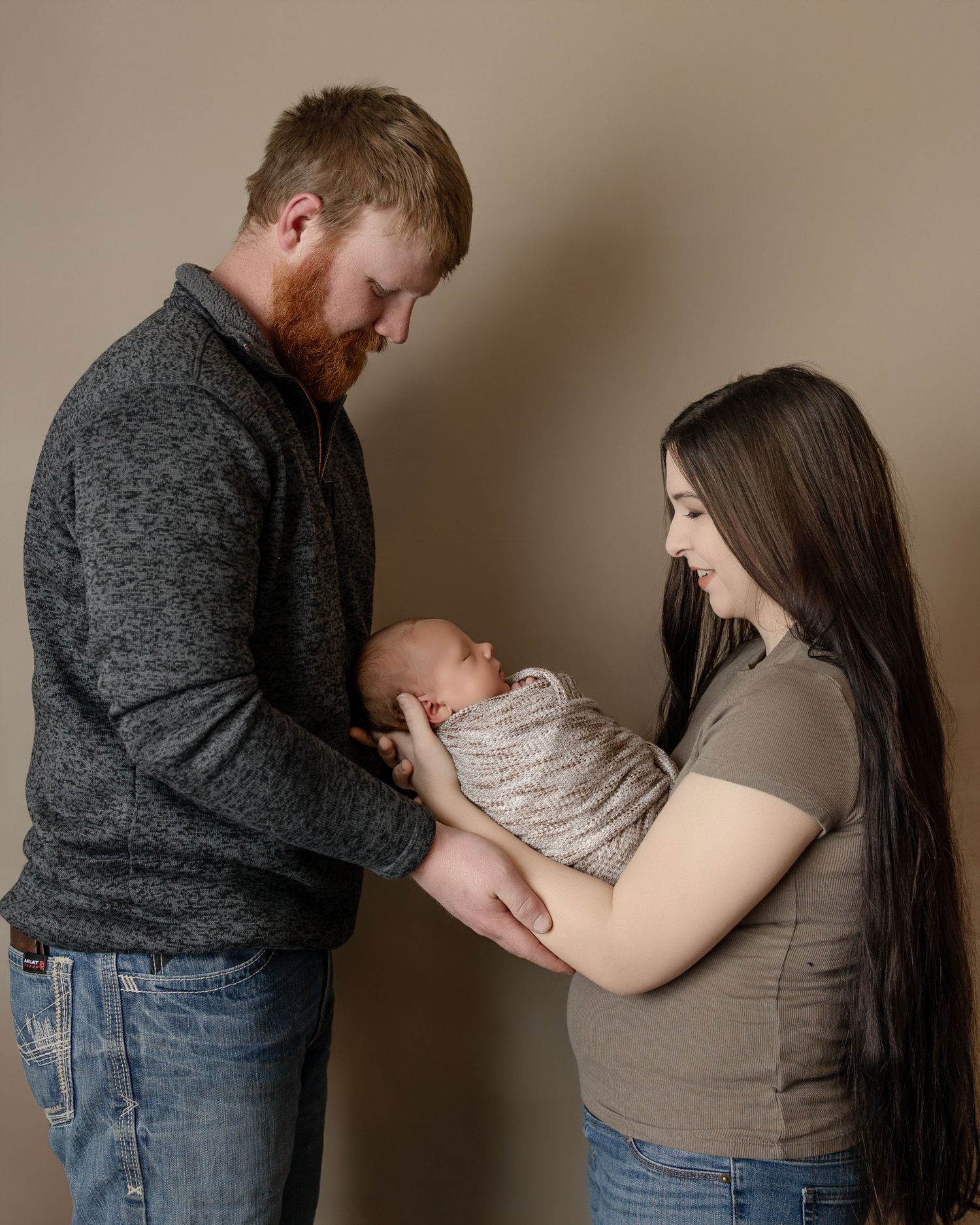 A couple gently holds their swaddled newborn baby in a neutral studio setting.