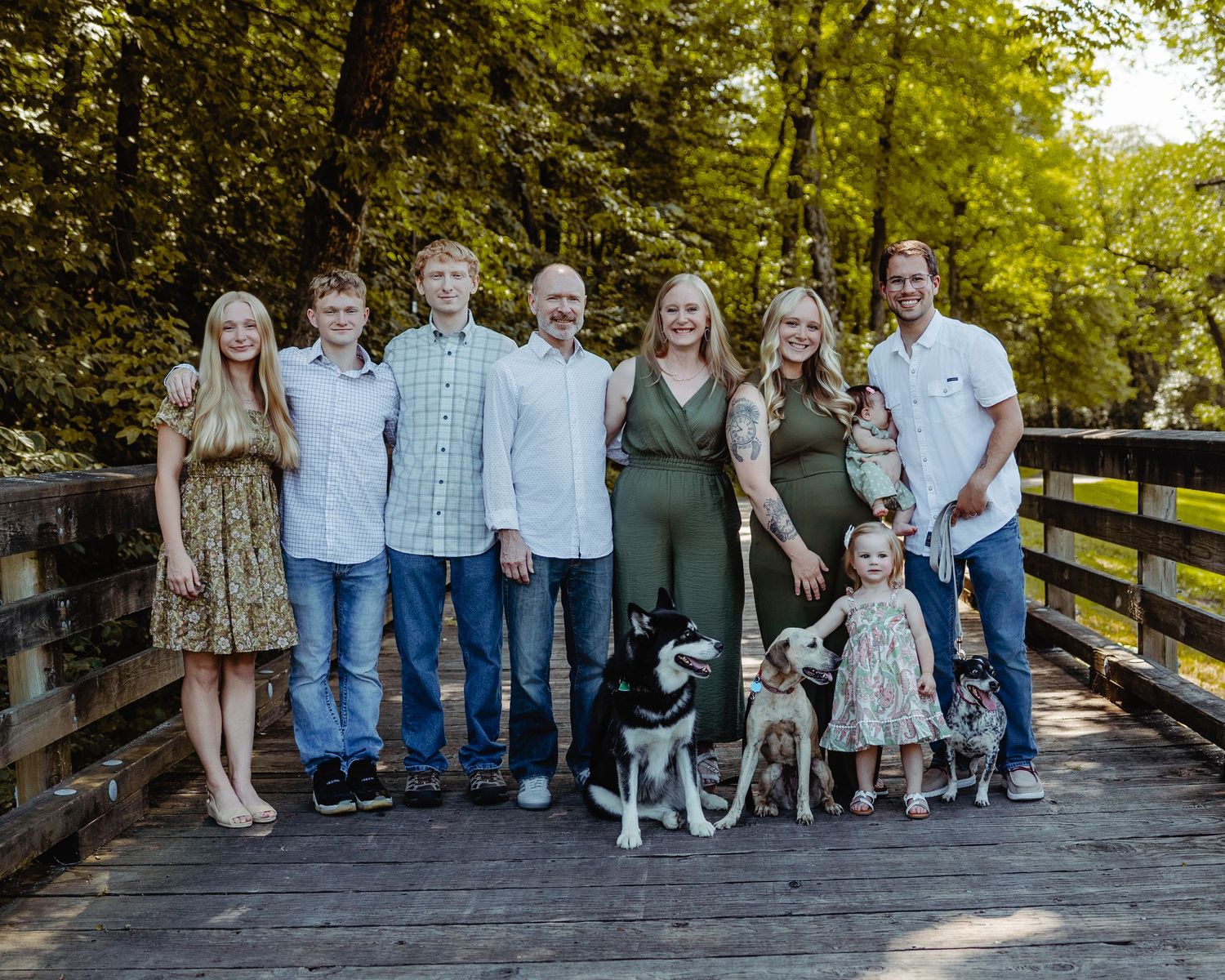 A group of people and their pets pose together on a wooden bridge surrounded by lush green trees in a park setting.