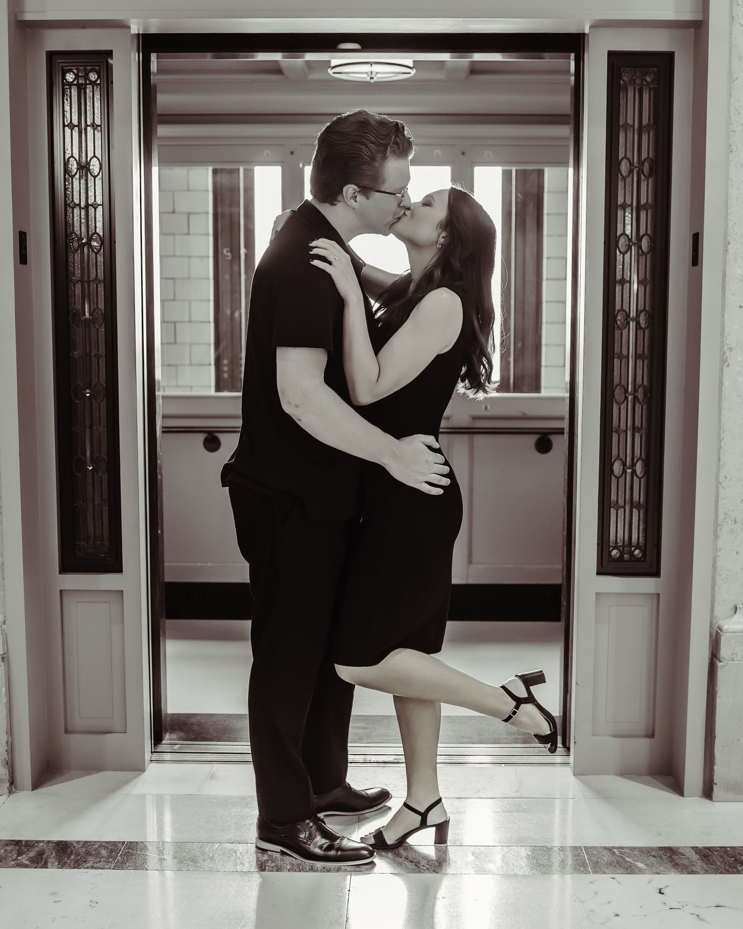 A romantic black and white portrait of a couple embracing in front of ornate art deco doors in an elegant hallway.