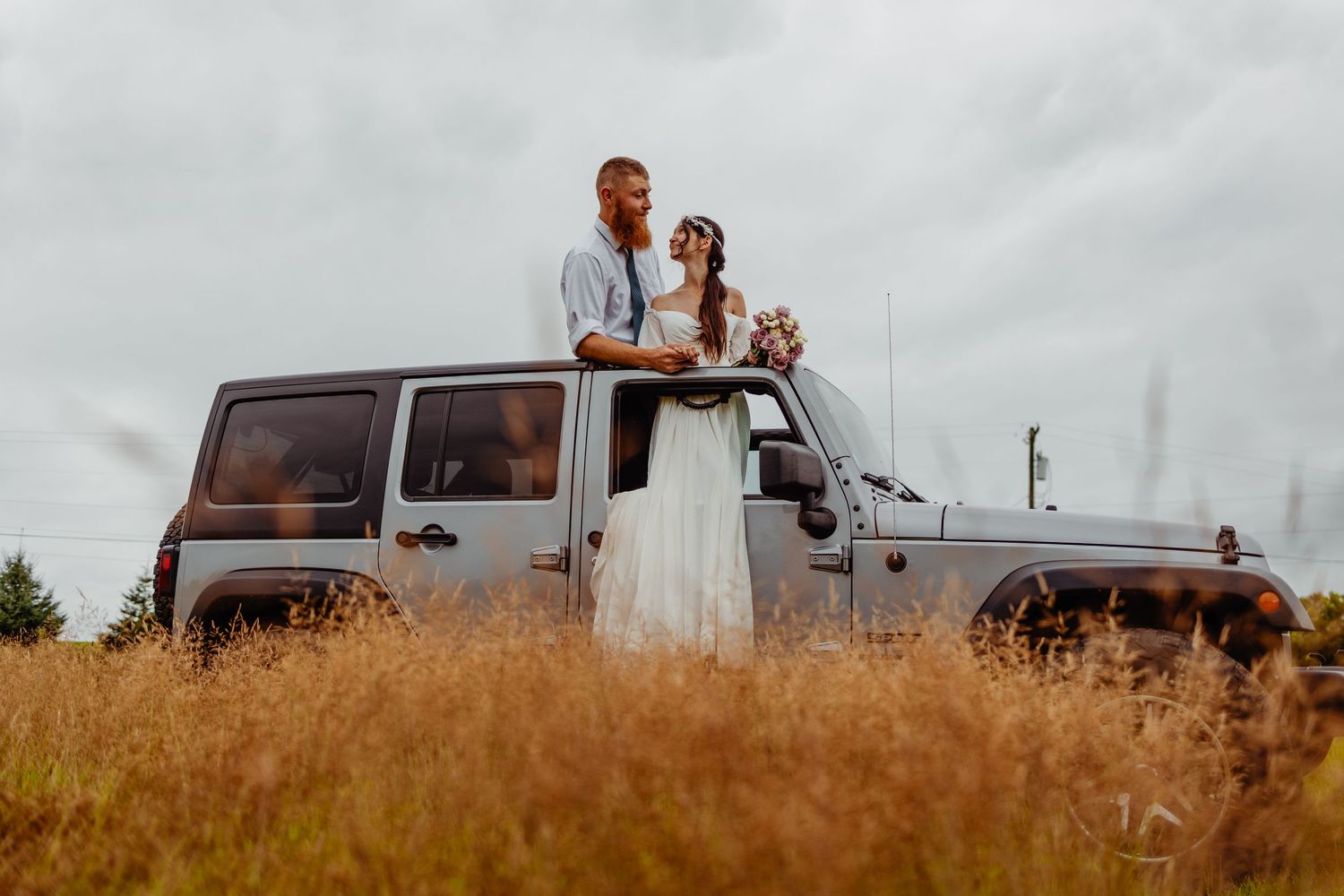 Newlyweds standing through sunroof of silver Jeep in golden field at sunset.