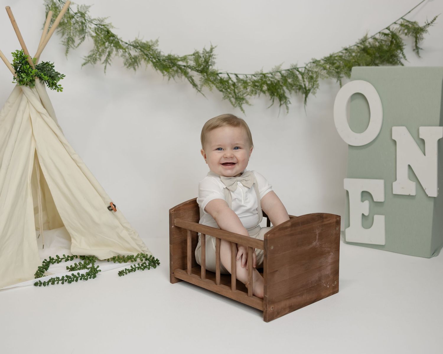 A smiling baby in a rustic wooden crate with a teepee and greenery garland in a minimalist nursery setting.