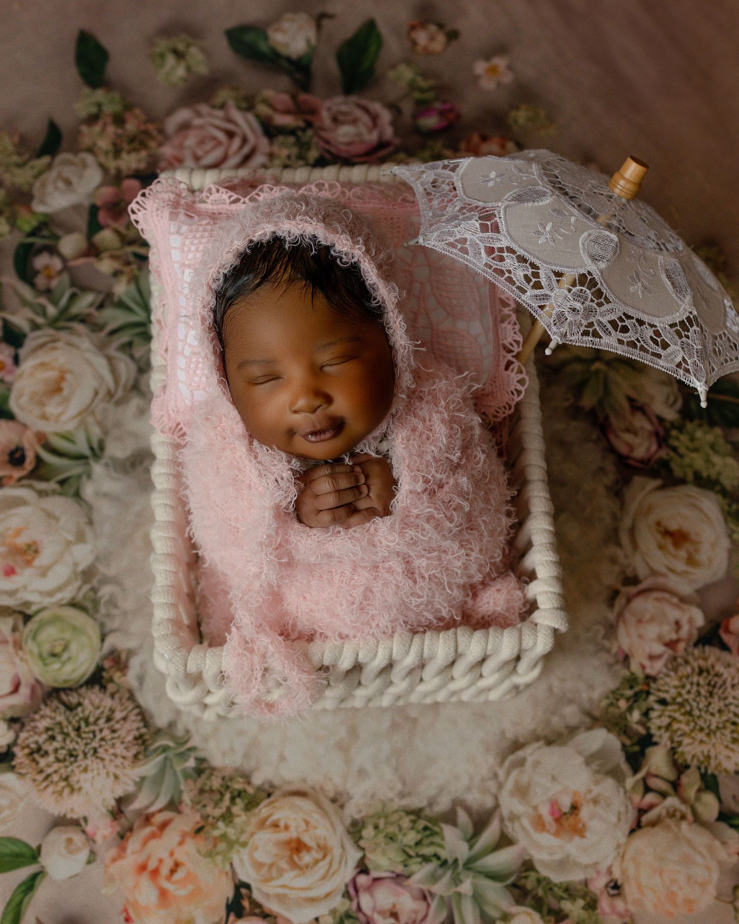 Newborn photography setup with pink and lace decorations surrounded by soft roses on a floral backdrop.