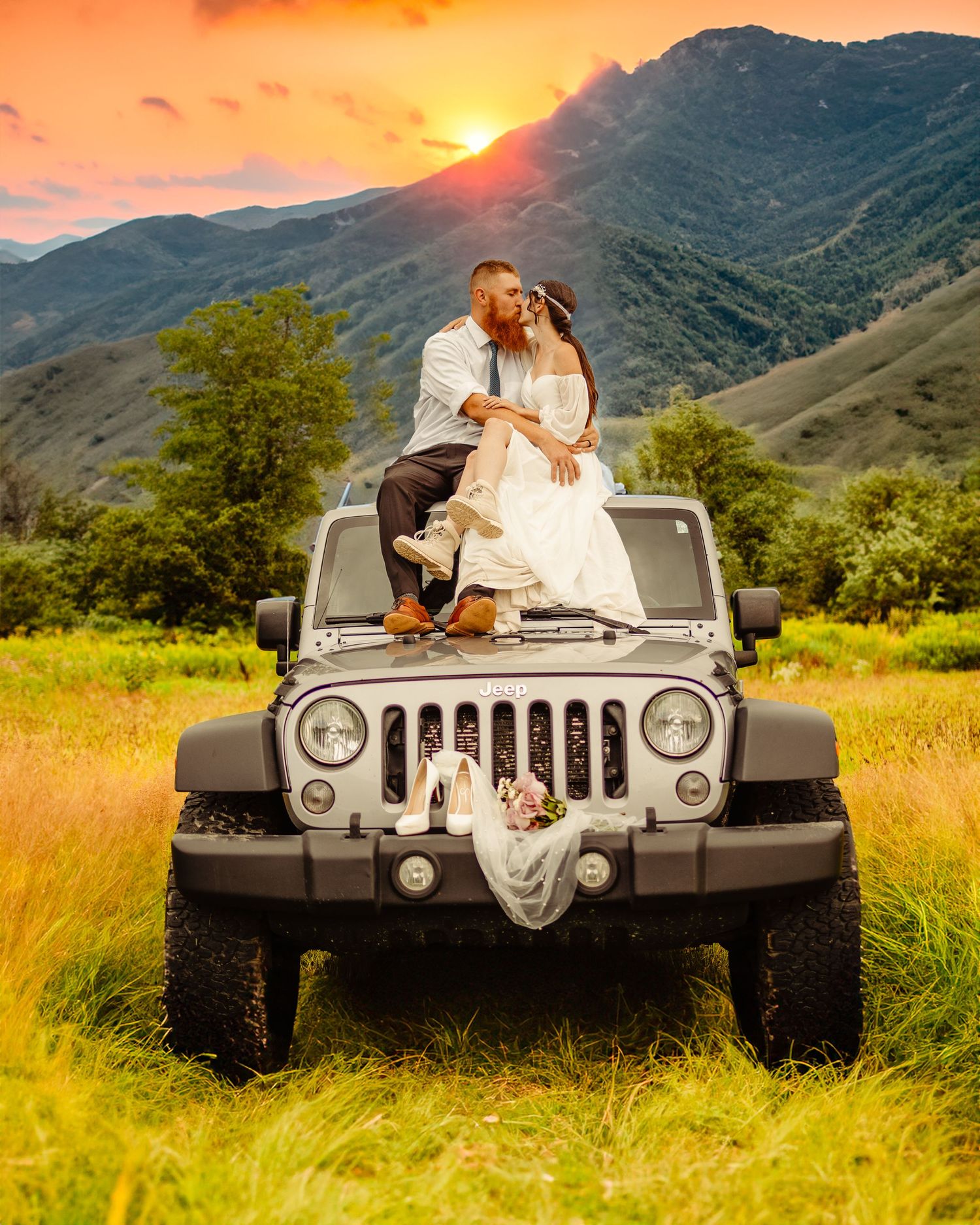 A couple sits atop a silver Jeep Wrangler during sunset in a mountainous landscape.