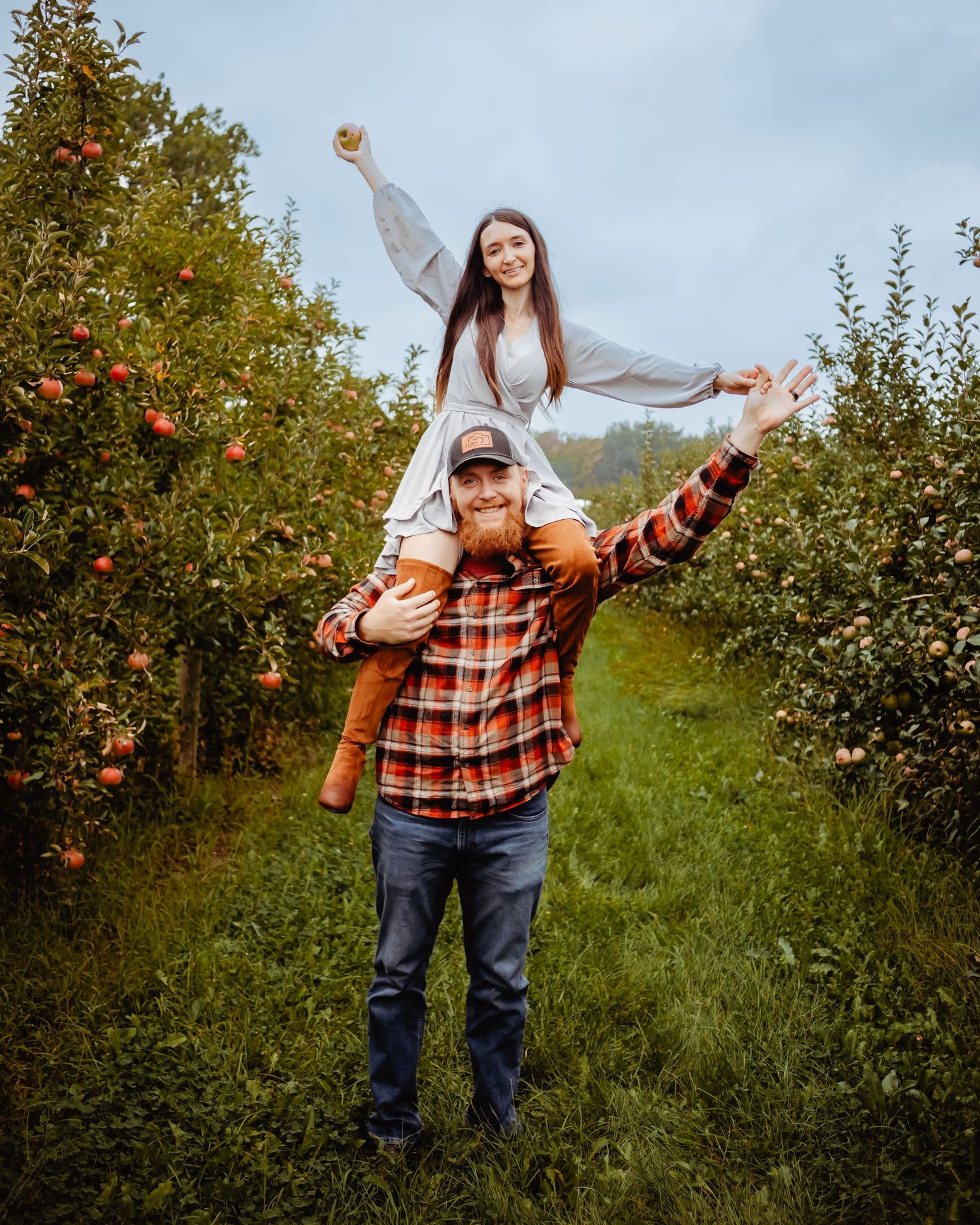 A playful couple poses in an apple orchard with one person sitting on shoulders against autumn trees.