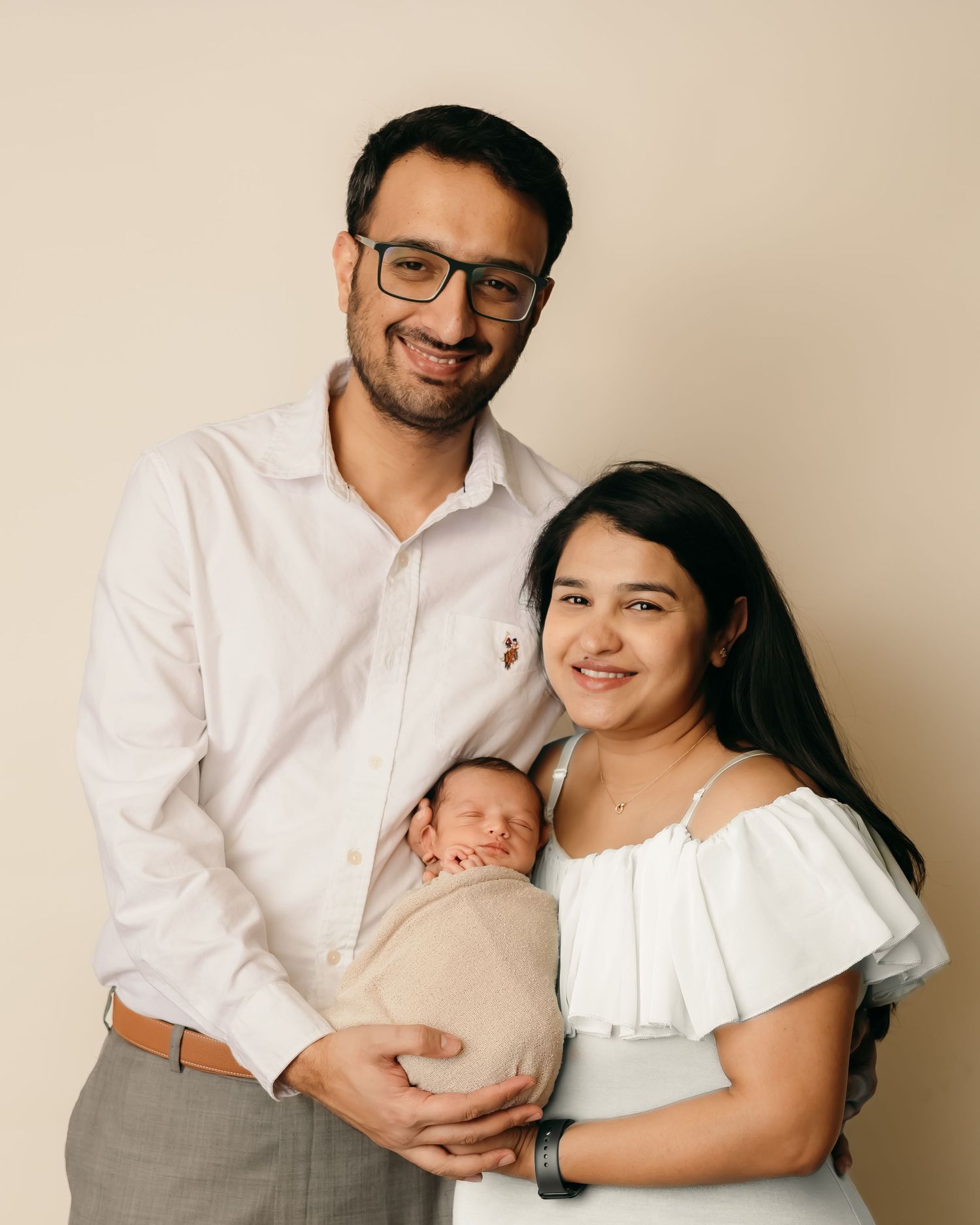 A couple in white garments hold their sleeping newborn baby against a neutral beige background in a studio setting.