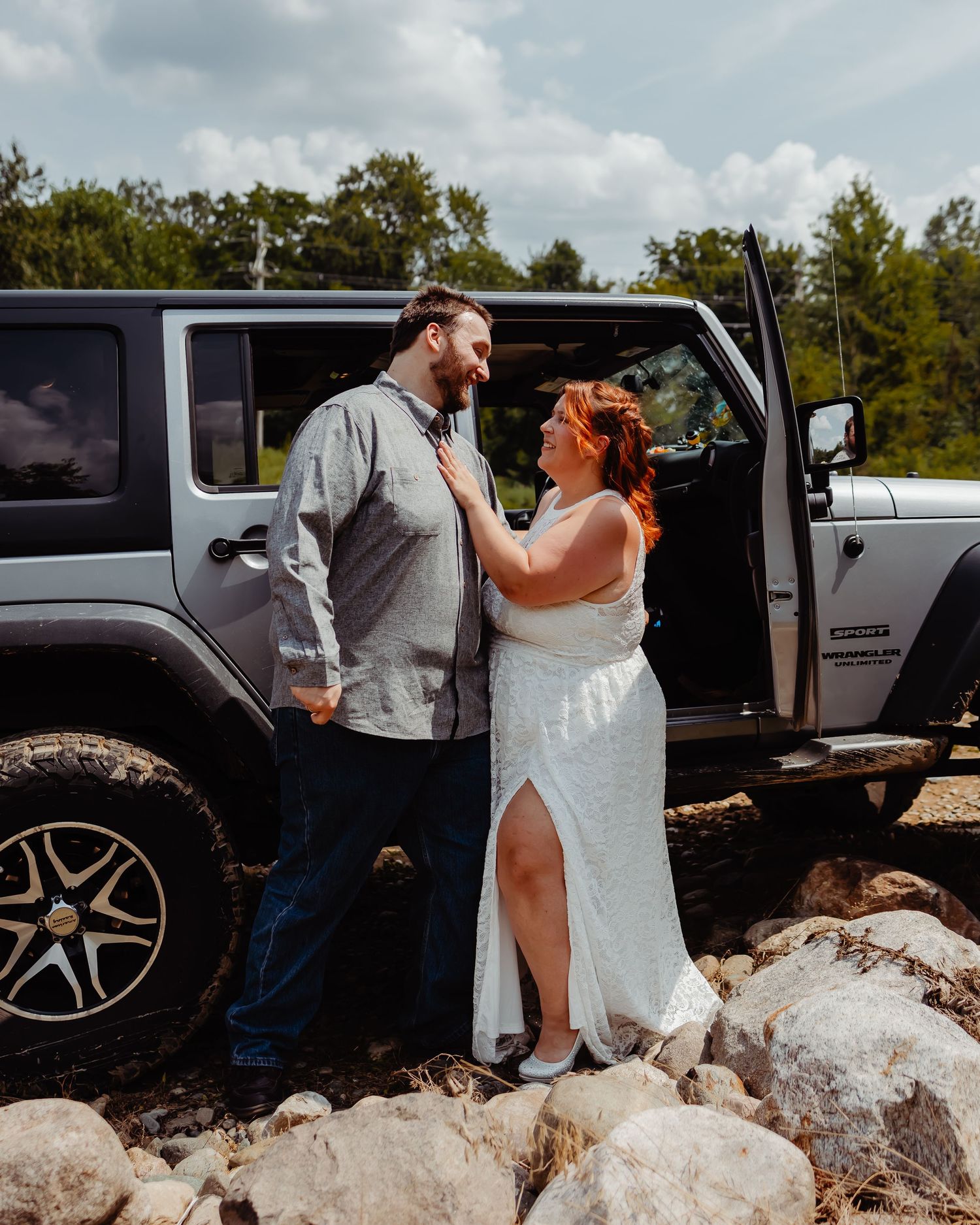 Newlyweds laugh together beside a black Jeep Wrangler on rocky terrain.
