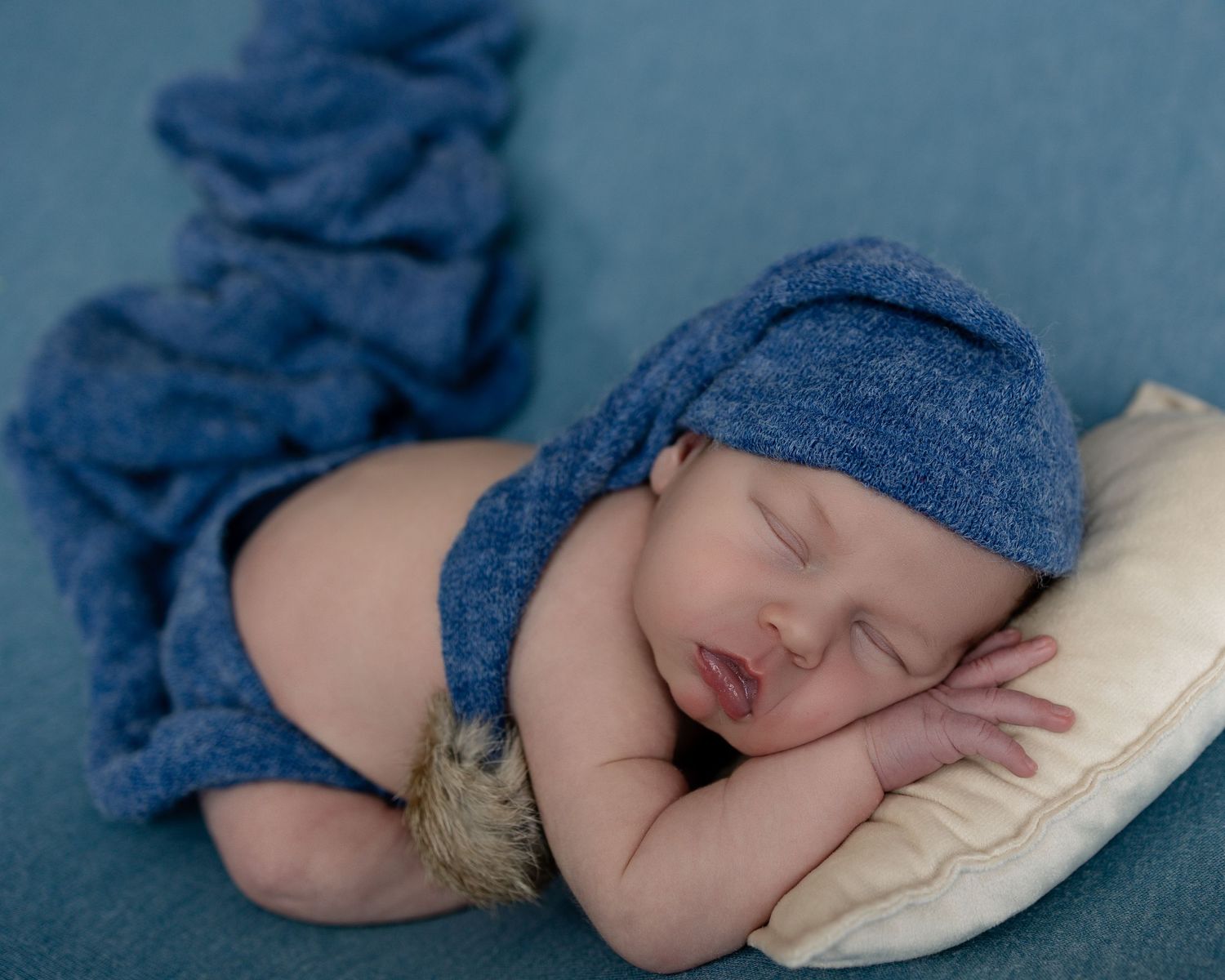 Sleeping newborn in blue knit hat and wrap rests peacefully on white pillow during professional photo session.