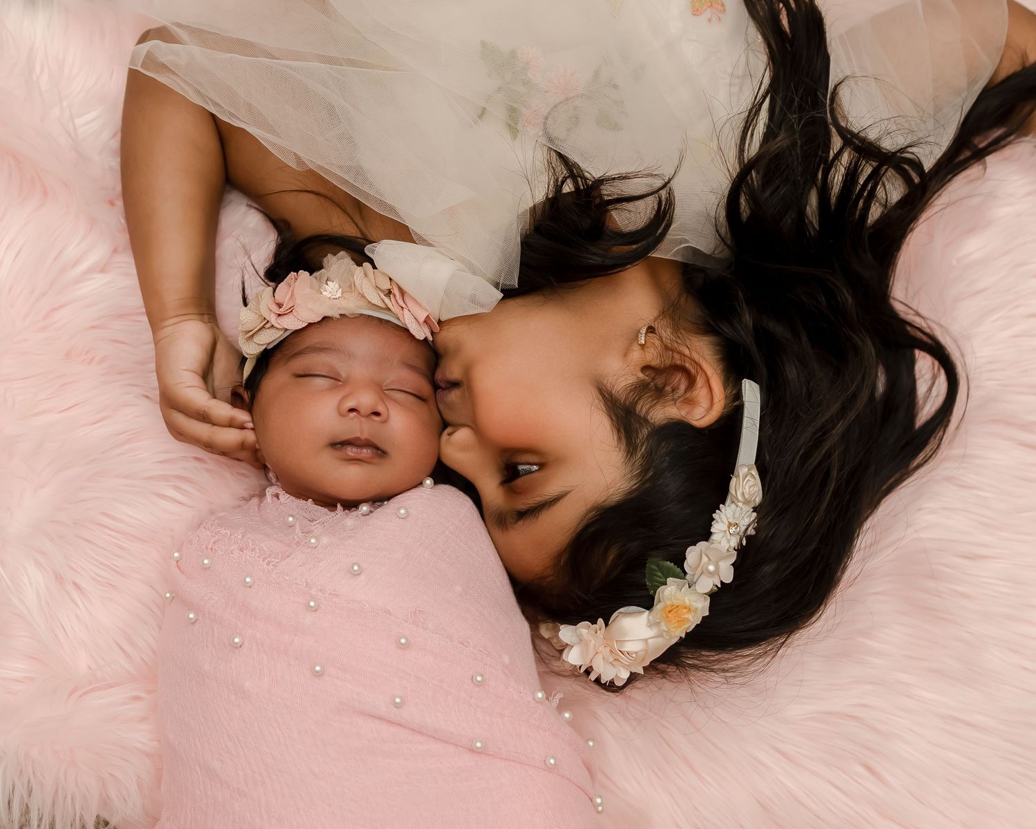 Parent and newborn baby lying together on soft pink fabric with white flower hair accessories.