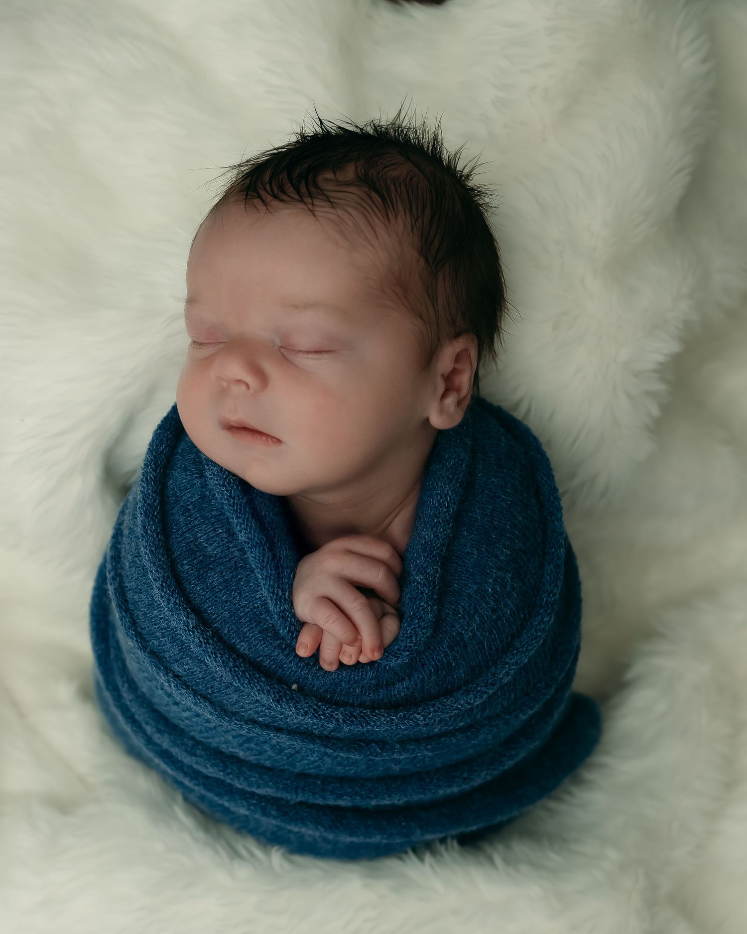 A sleeping newborn wrapped in a blue blanket rests peacefully on soft white fur.