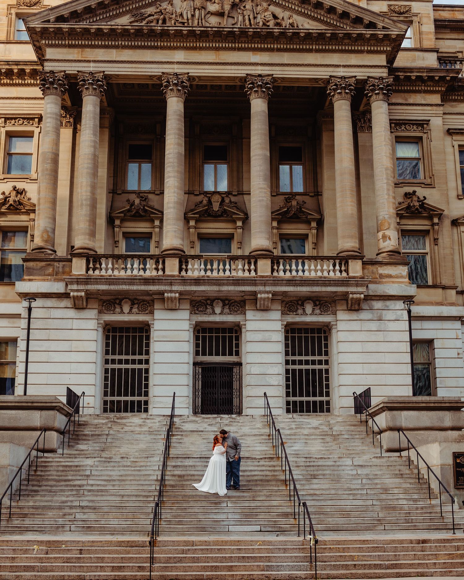 A couple poses on grand stone steps of a classical government building with ornate columns.