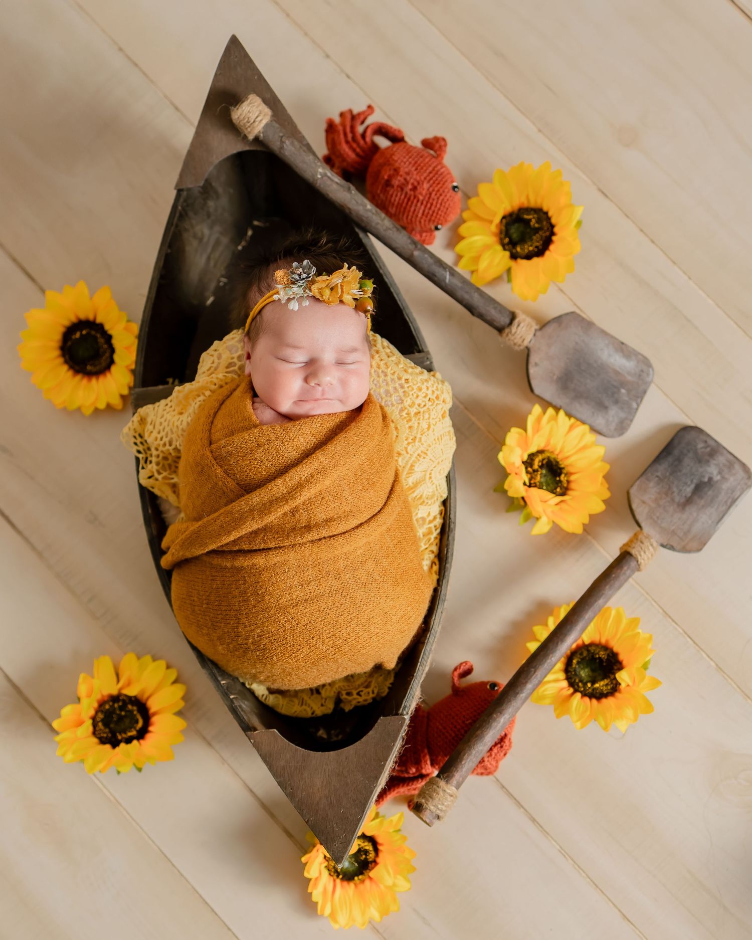 Overhead view of an infant swaddled in yellow fabric lying in a small boat surrounded by bright sunflowers and rustic props.