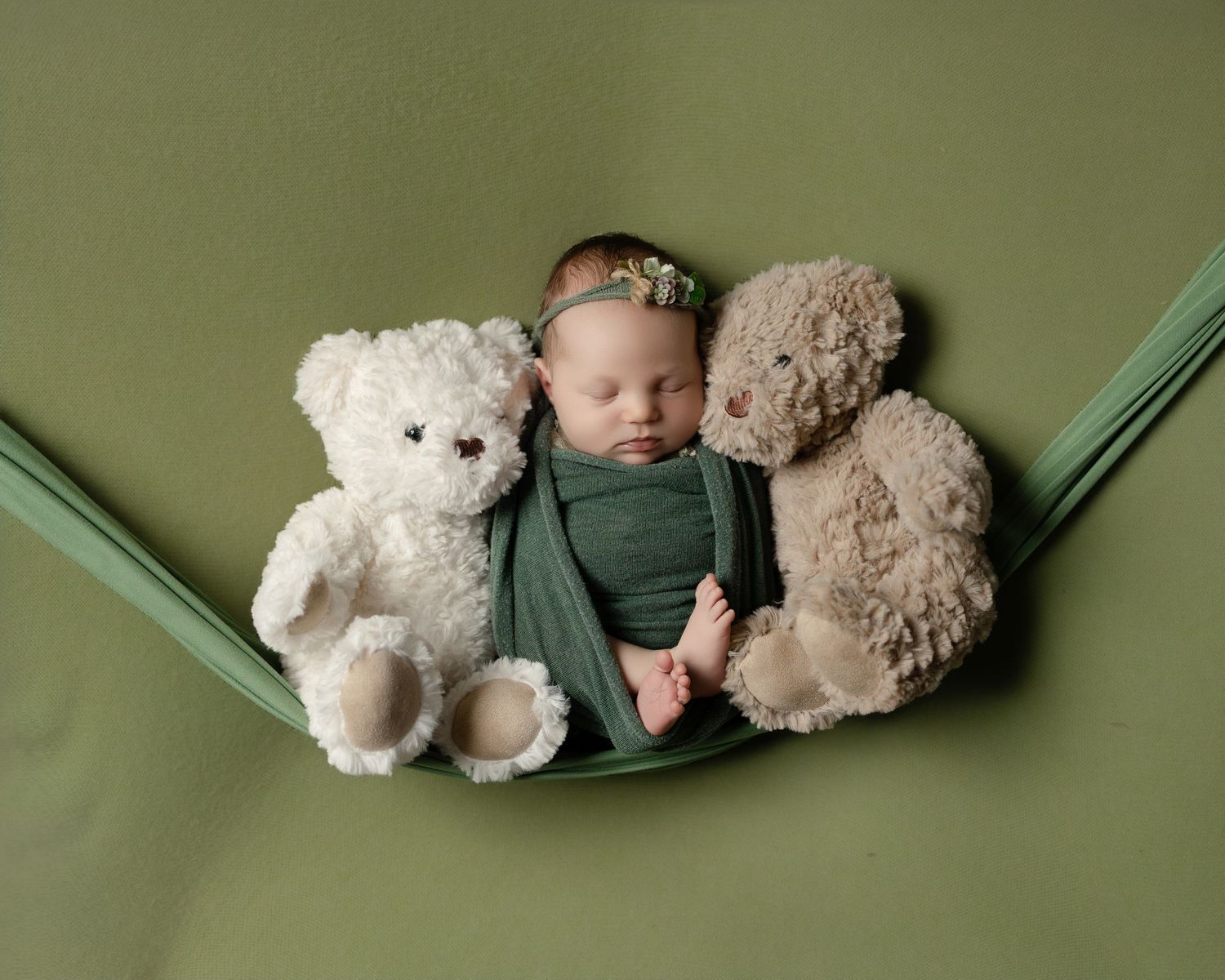 Newborn baby sleeping in green hammock wrap between two teddy bears on green background.