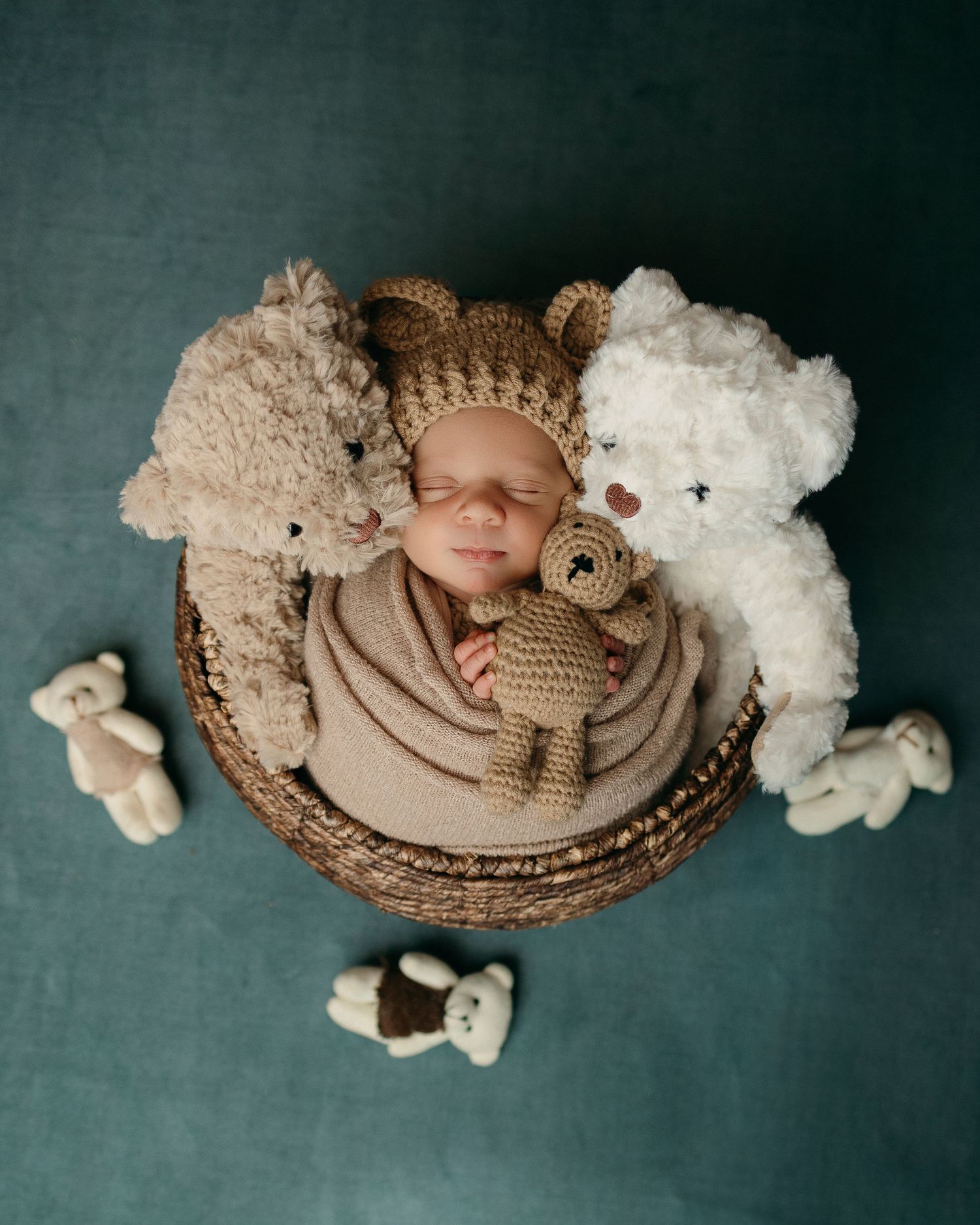 Overhead view of sleeping baby in brown knitted bear hat surrounded by plush teddy bears in a round basket.