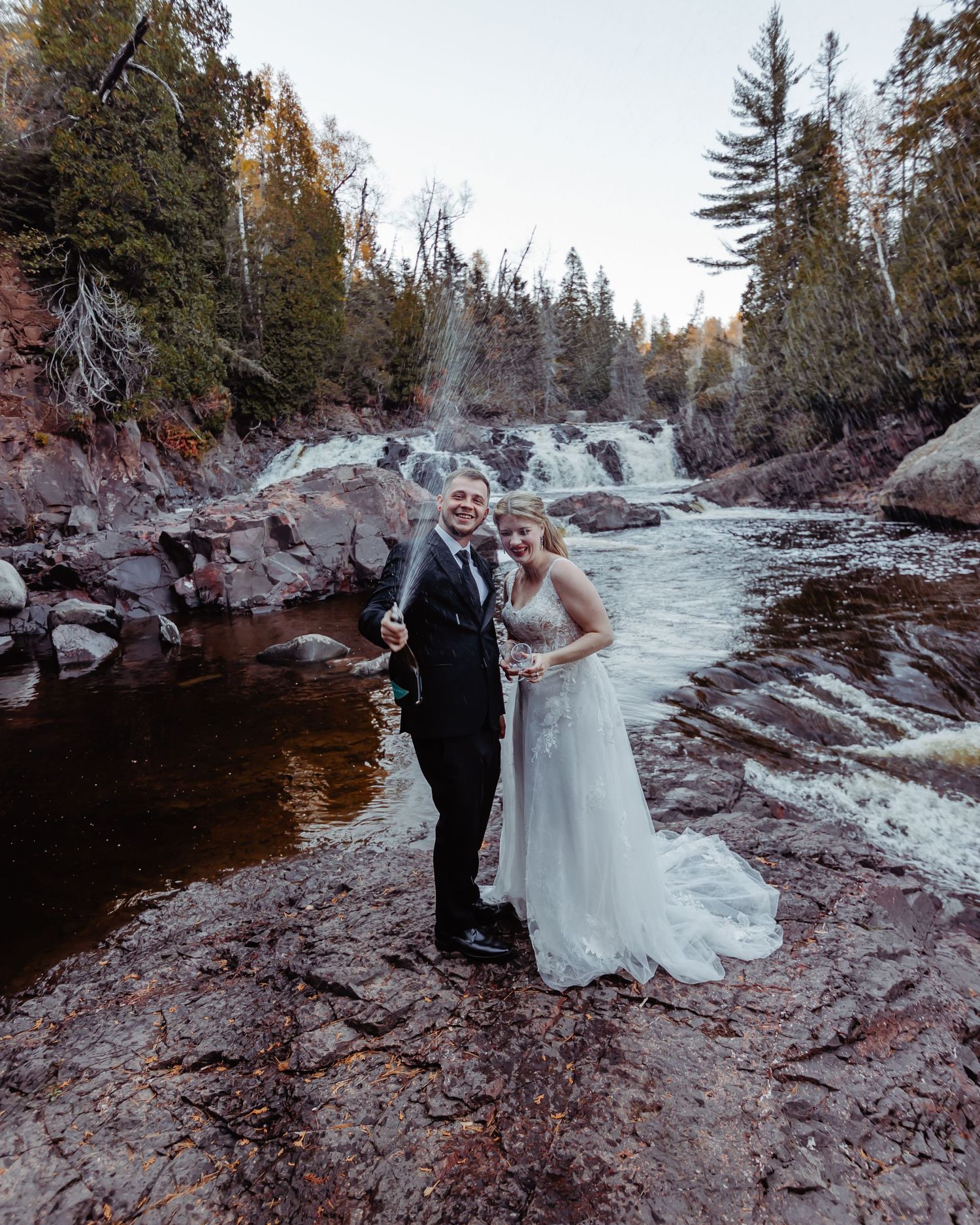 An elopement couple stands on red rocks near cascading waterfalls in a forested setting.