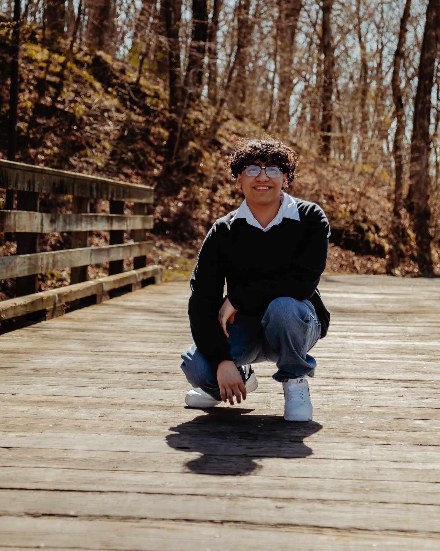 Someone in a black sweater and glasses crouches on a wooden boardwalk path through an autumn forest.
