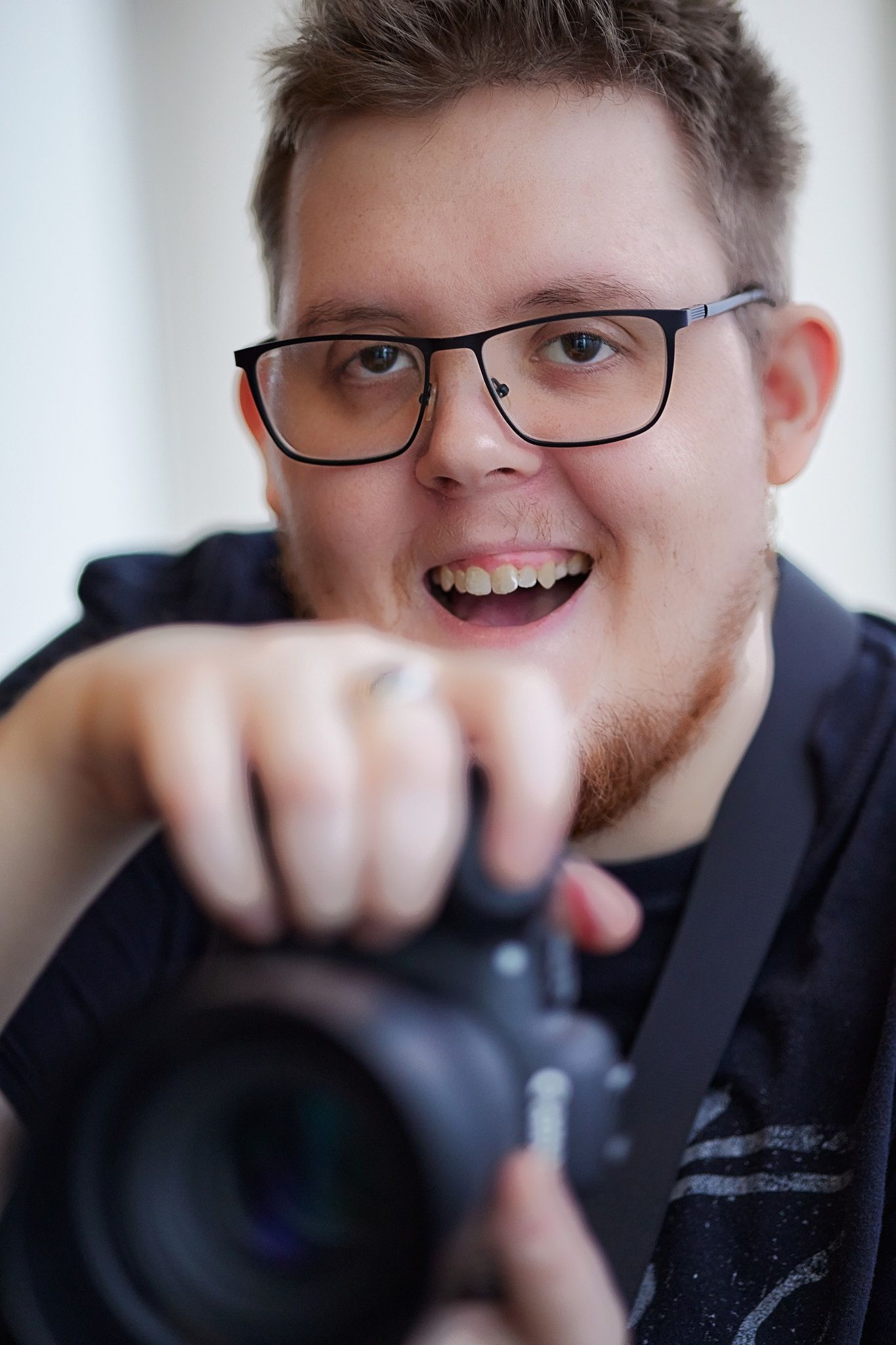Energetic photographer wearing black glasses and a navy shirt holds a professional camera with an excited expression.