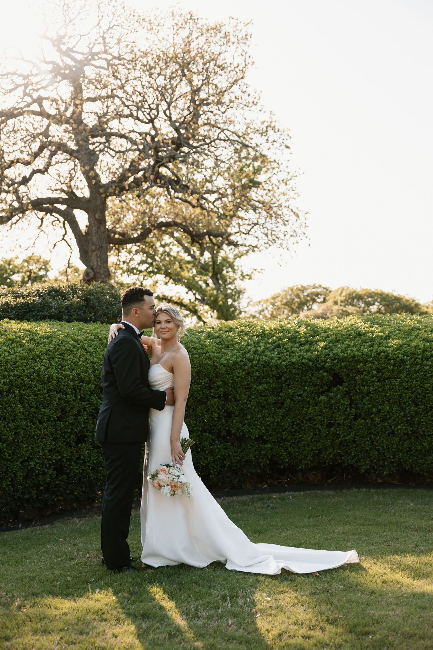 Newlyweds share an intimate moment at sunset in a garden setting with tall hedges and a majestic tree in the background.