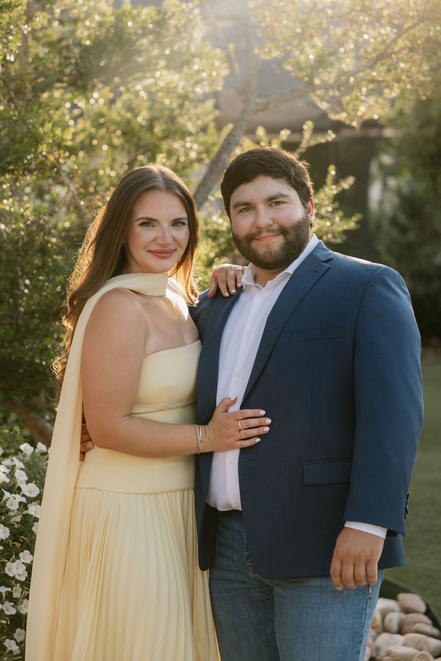 Couple in formal attire embraces in an outdoor garden setting during golden hour with soft natural lighting.