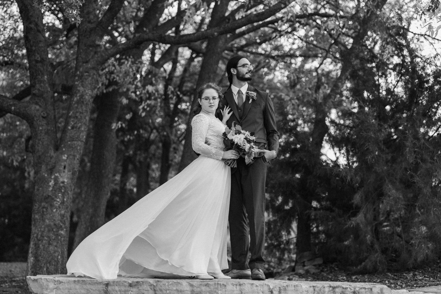A black and white wedding photo captures a couple in formal attire standing among trees with the bride's dress flowing in the wind.