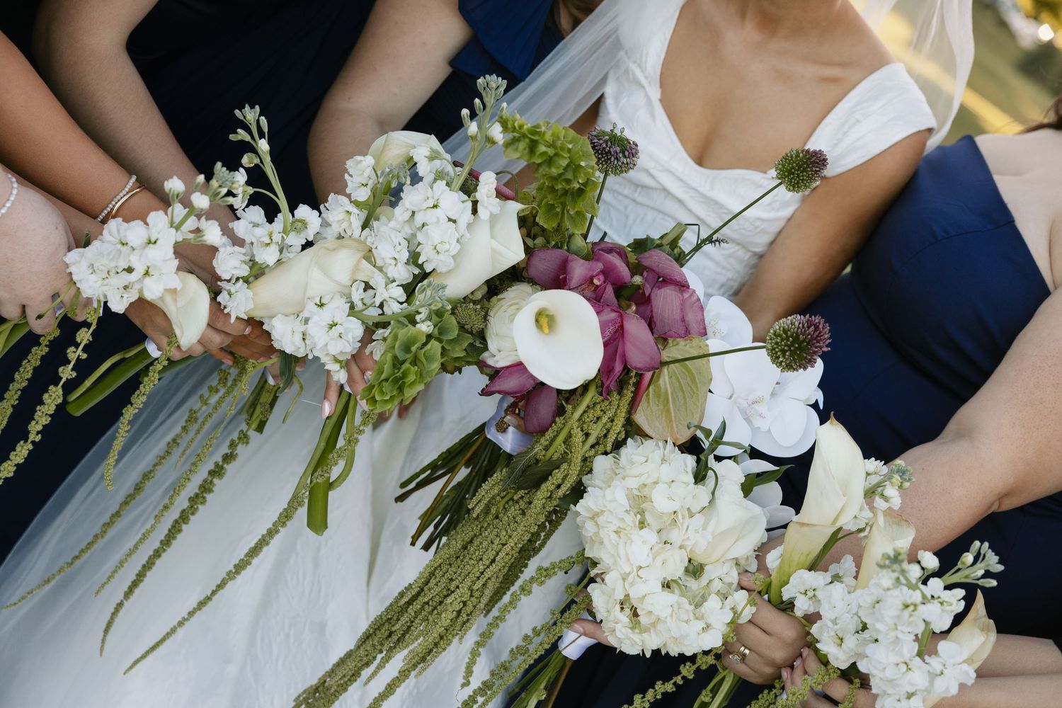 A wedding party holds elegant bouquets featuring white hydrangeas, roses and cascading greenery against navy attire.
