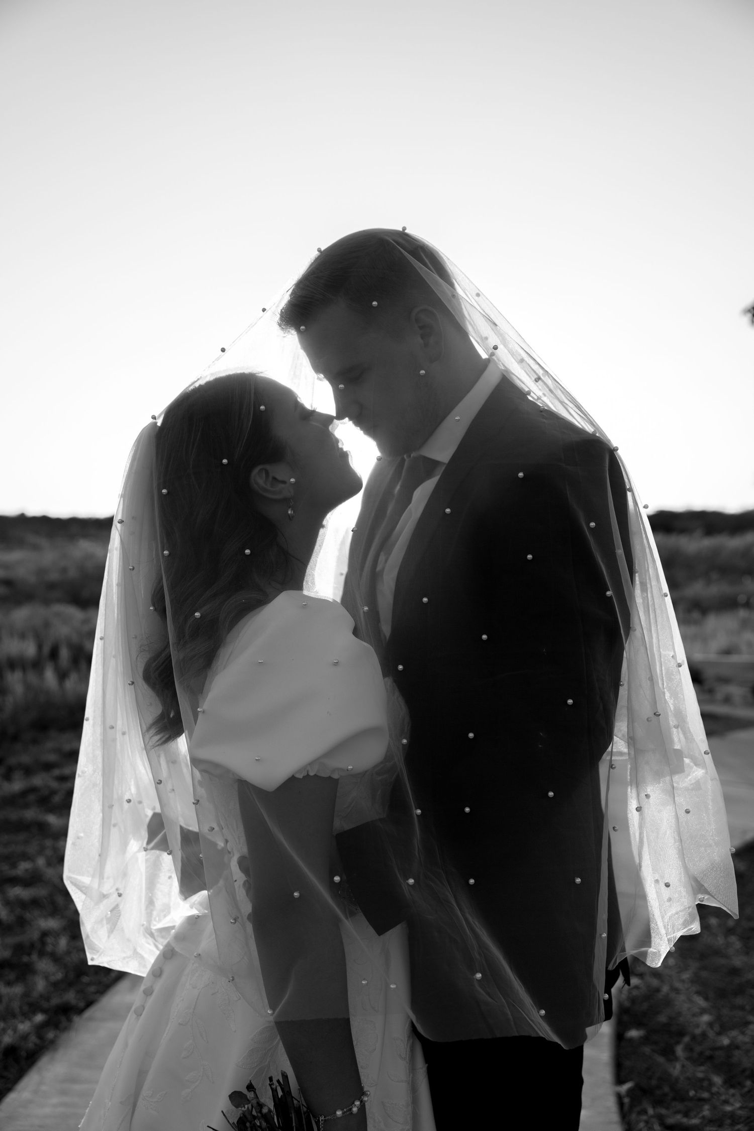 A romantic black and white wedding photo silhouette of a bride and groom standing close together under a flowing veil.