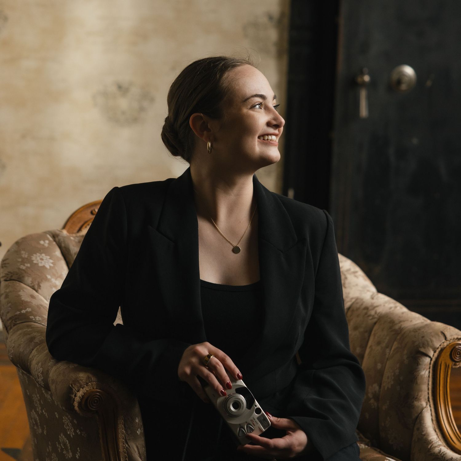 Person in black blazer sits on vintage chair while looking up with natural window light creating moody atmosphere.