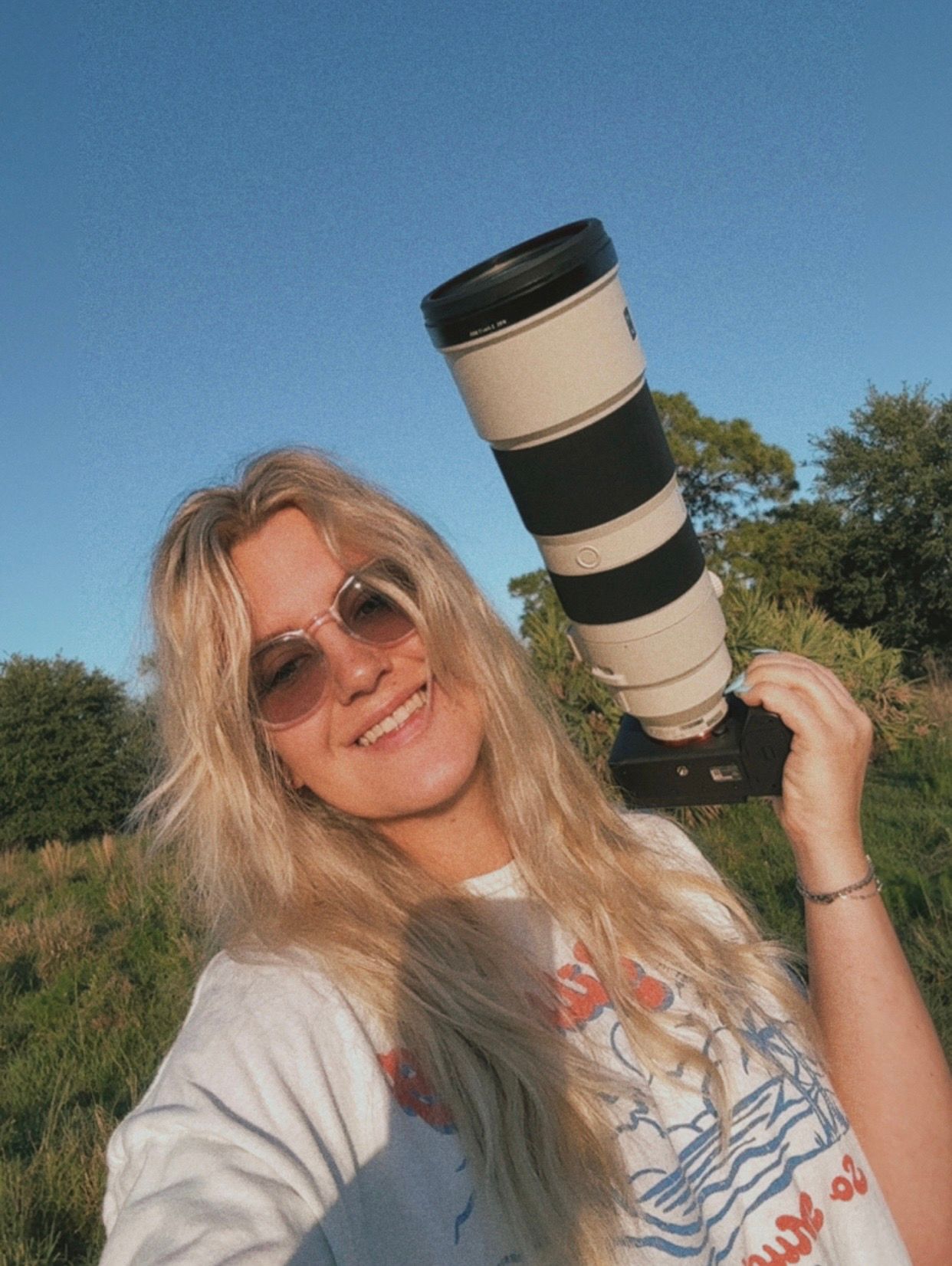 Smiling blonde woman holding a large Canon telephoto lens camera outdoors on a sunny day.