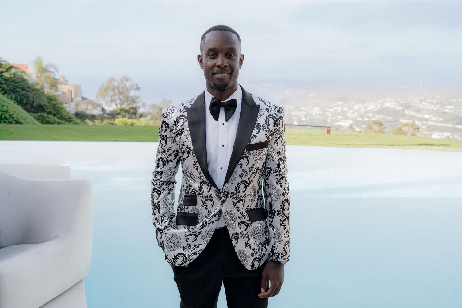 Man in floral tuxedo jacket and bow tie posing outdoors with scenic city view backdrop.