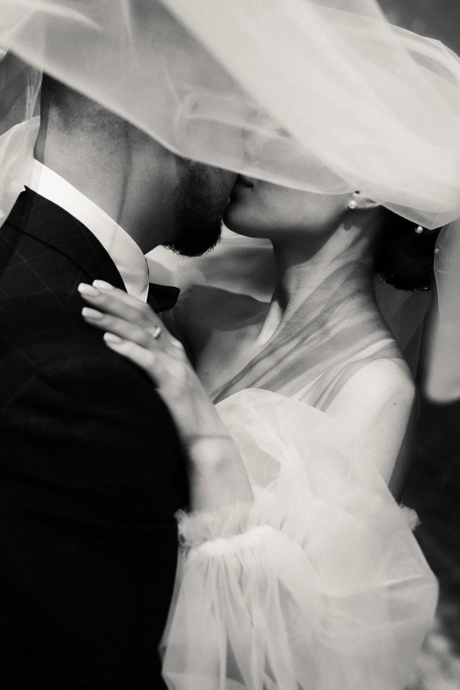 Black and white photo of bride and groom kissing under wedding veil, romantic couple portrait.