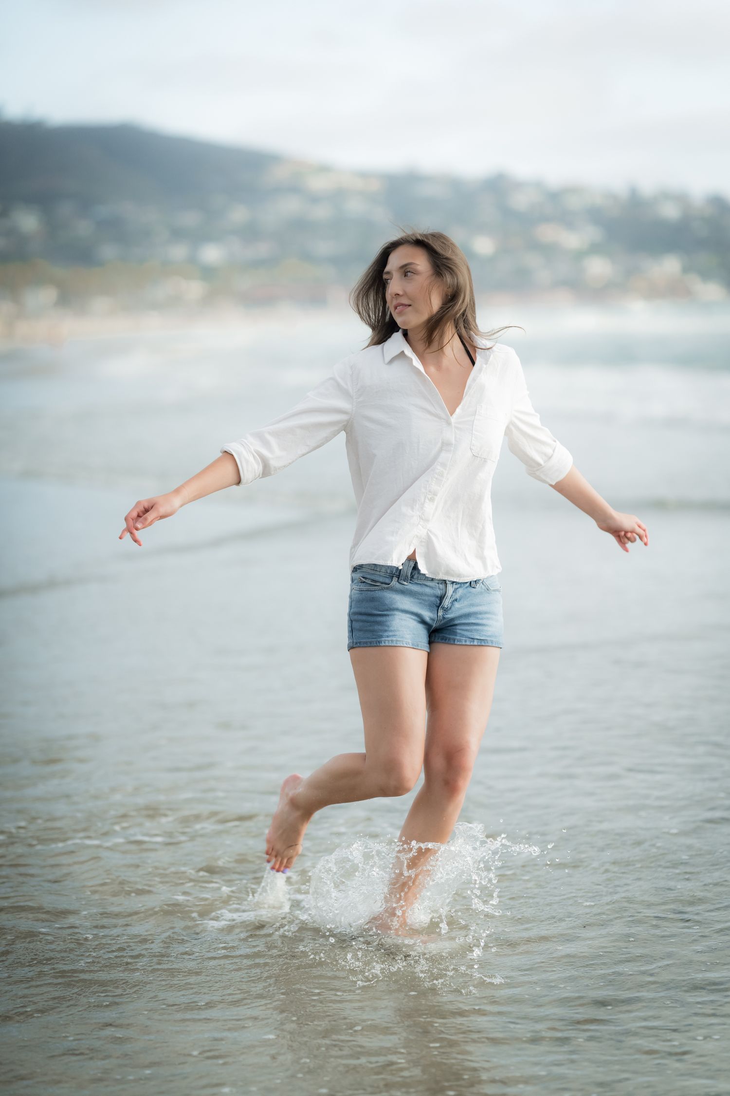 Woman in white shirt and denim shorts splashing in ocean waves at the beach on a cloudy day.