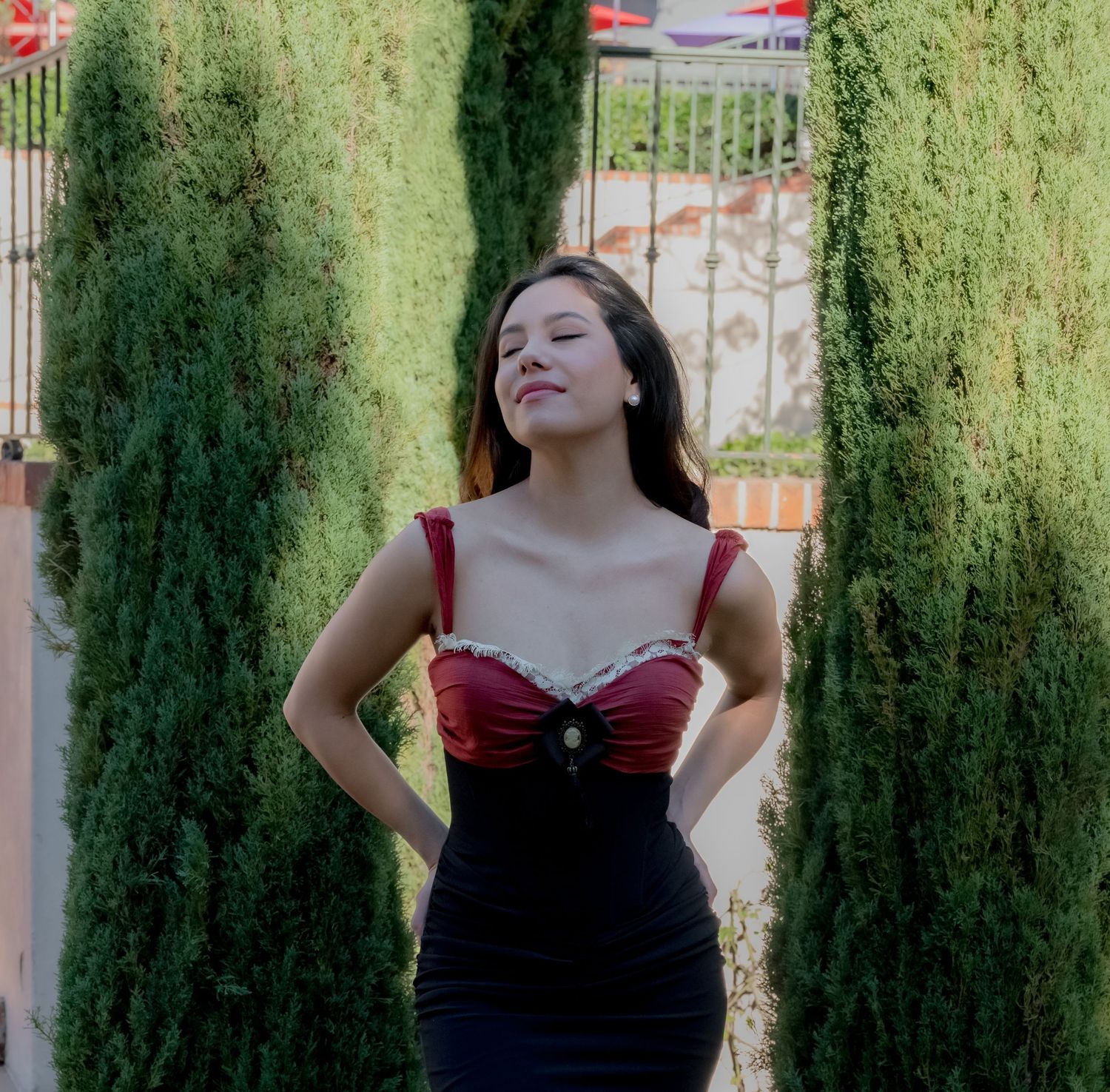 A young woman in a red and black dress poses confidently between tall cypress trees in an outdoor garden setting.