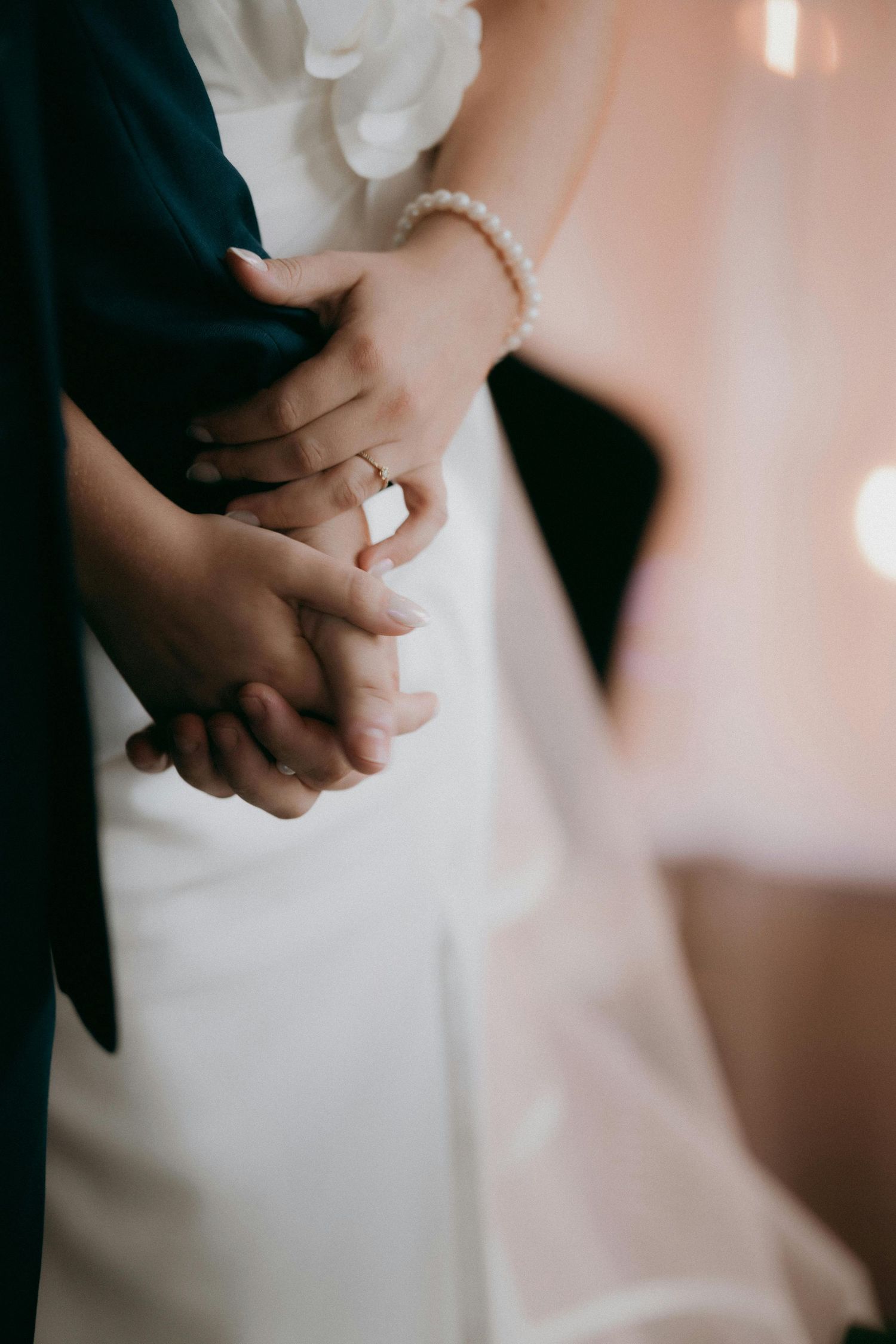 Bride and groom holding hands, showcasing a pearl bracelet and engagement ring, with wedding attire visible.