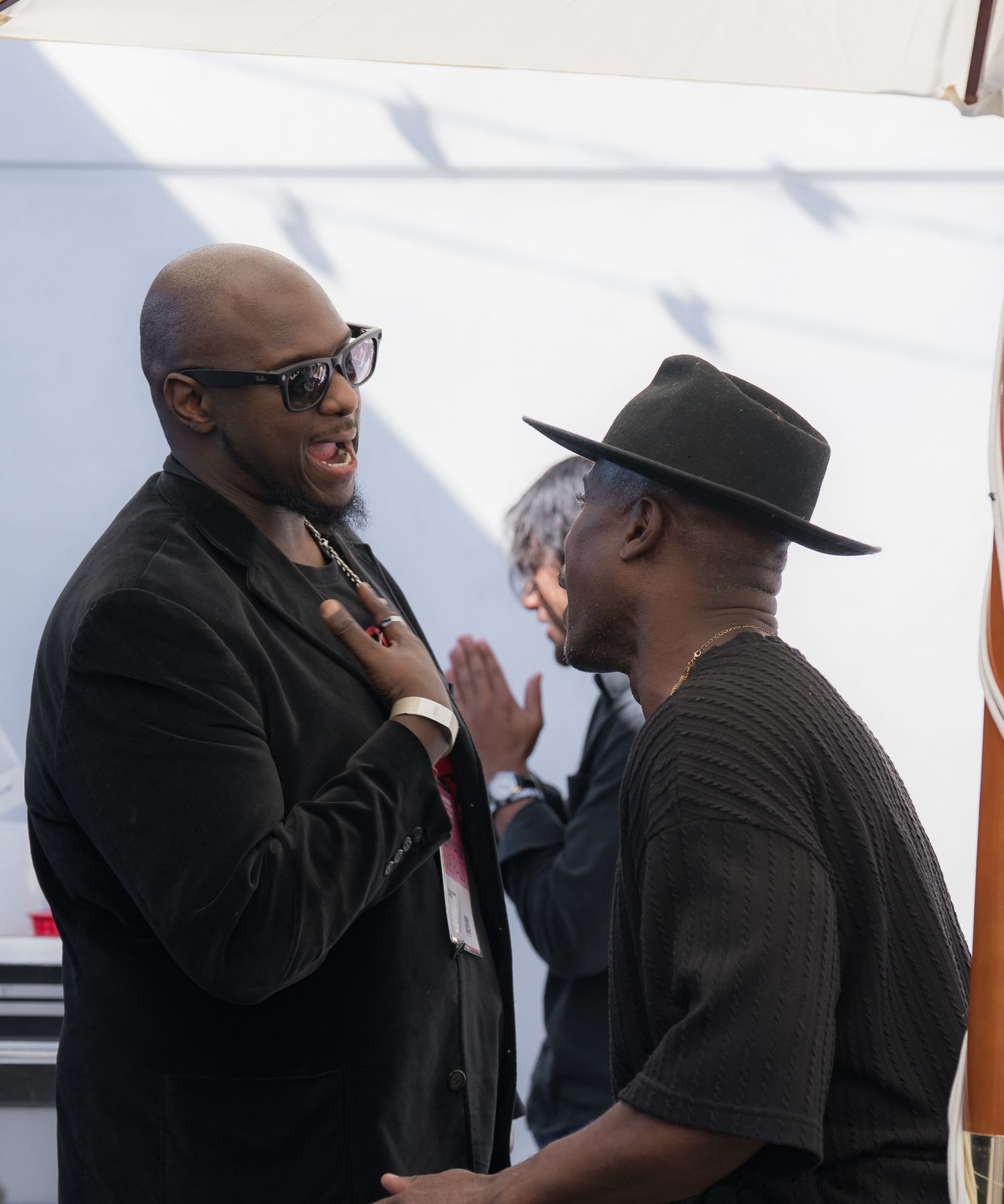 Two men in black attire engage in conversation outdoors, one wearing sunglasses and one in a wide-brim hat.