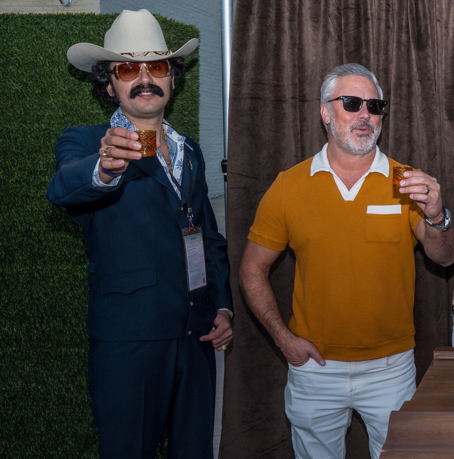 Two men in retro outfits holding drinks, one in a cowboy hat and suit, the other in an orange polo shirt.