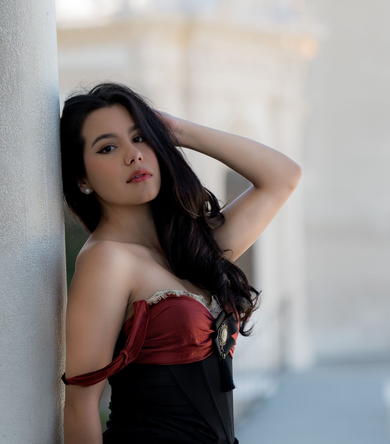 Young woman in red strapless corset top leaning against a column with hand in dark hair.