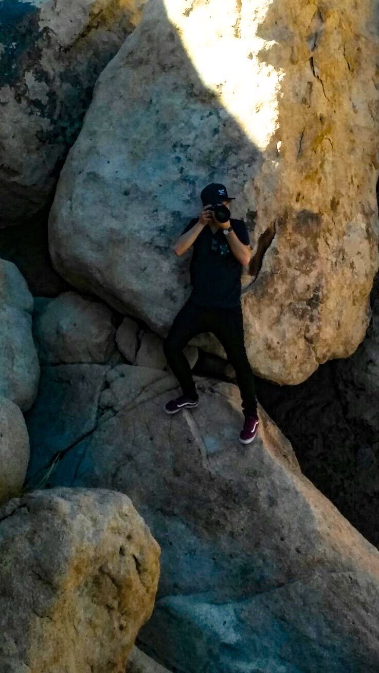 Photographer in black outfit and cap taking photos while standing between large boulders.