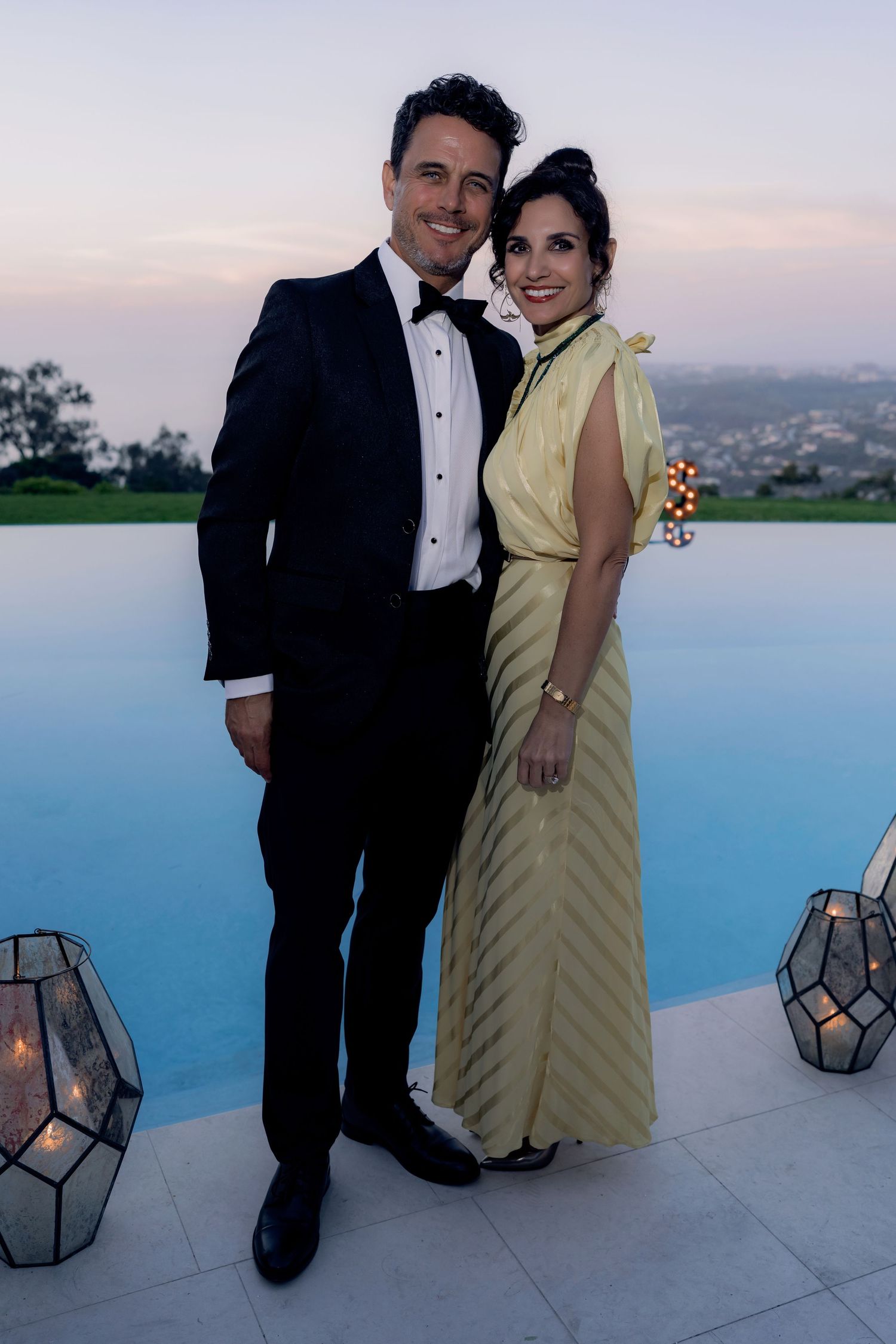 Elegant couple posing by infinity pool at dusk, man in tuxedo and woman in yellow maxi dress at formal event.
