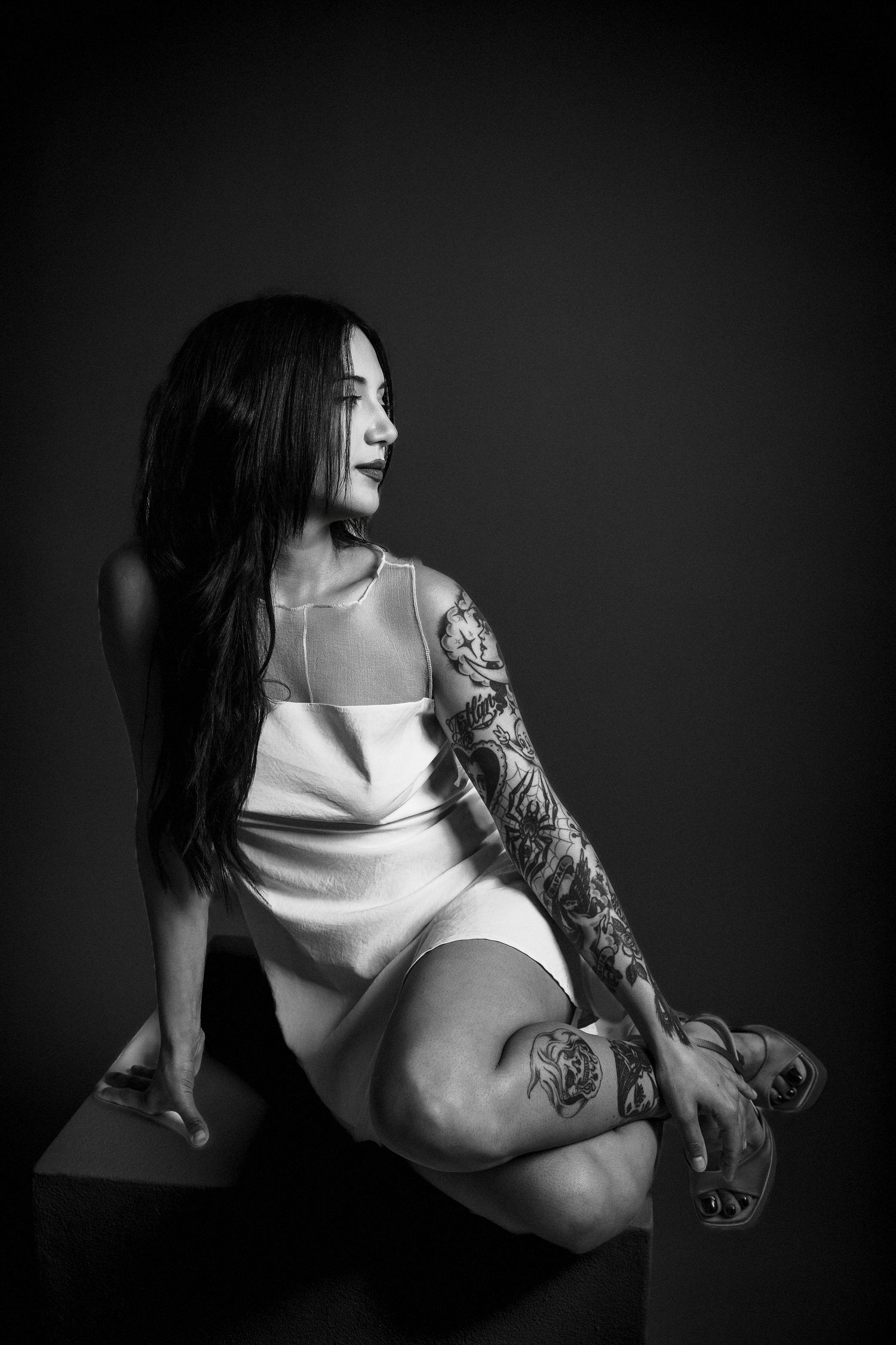 Black and white portrait of a tattooed woman in a white dress sitting gracefully on a cube against a dark background.