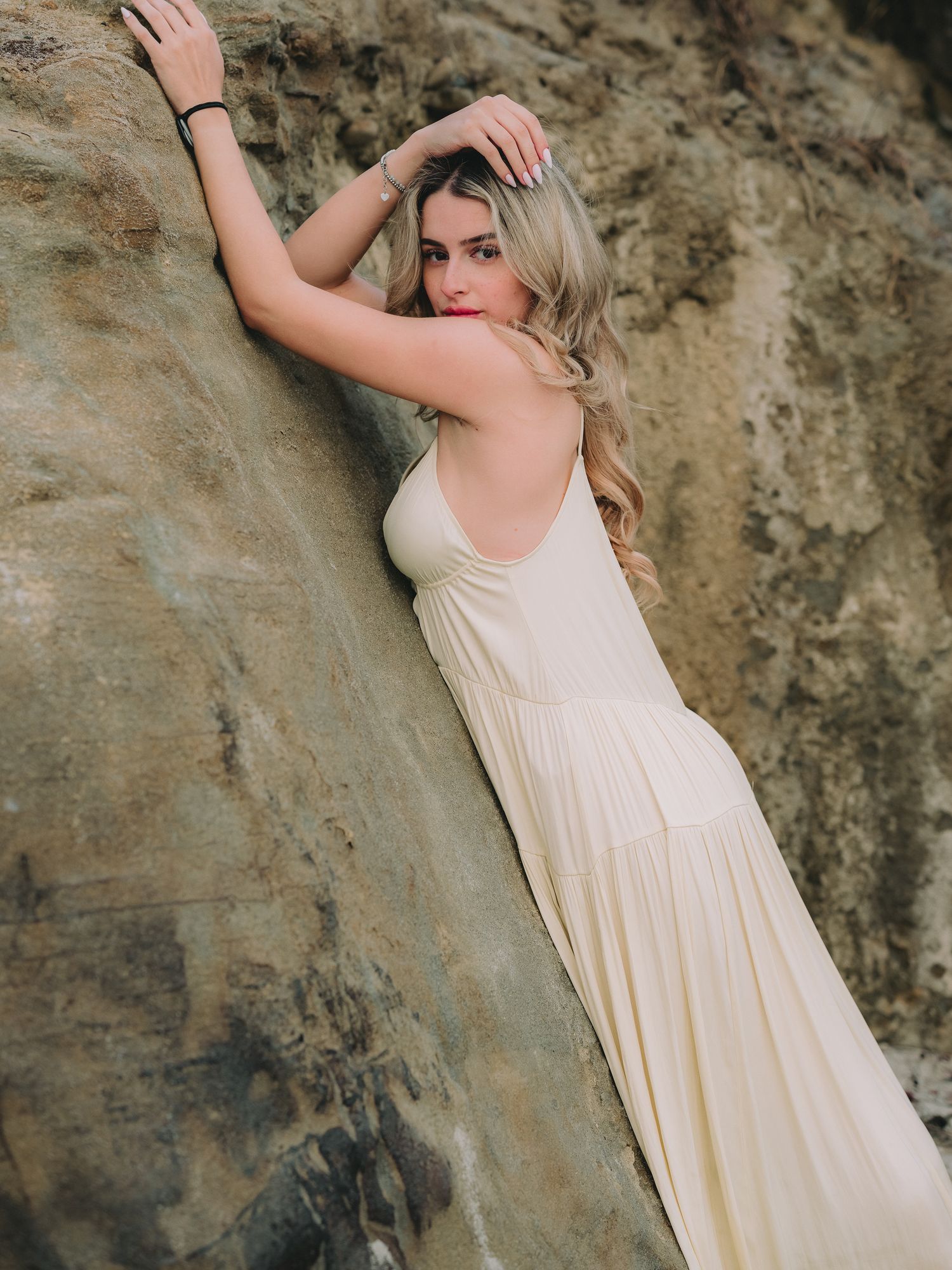 A blonde woman in a cream maxi dress leans gracefully against a rocky cliff in an outdoor fashion photo shoot.
