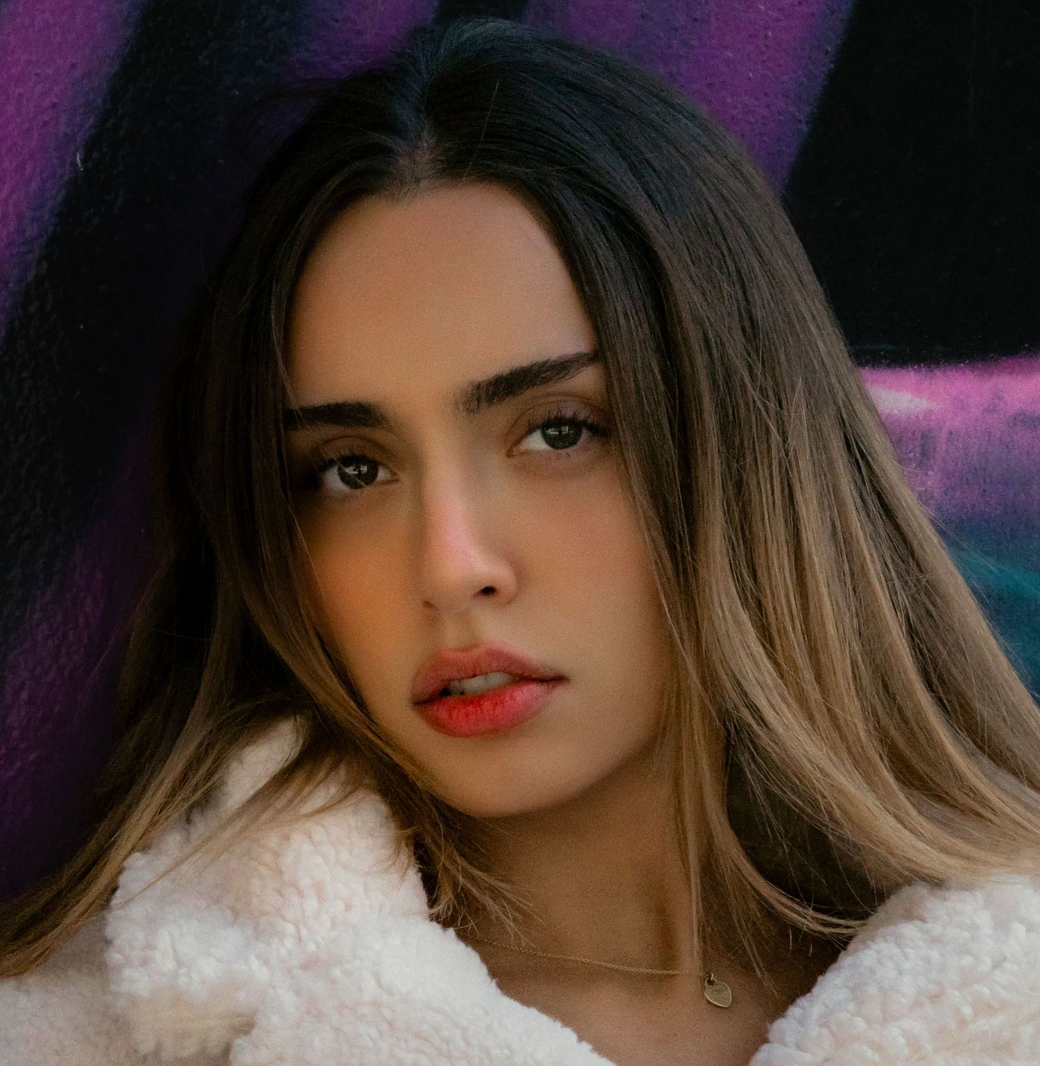 Young woman with ombre hair in white fluffy coat posing against colorful graffiti wall.