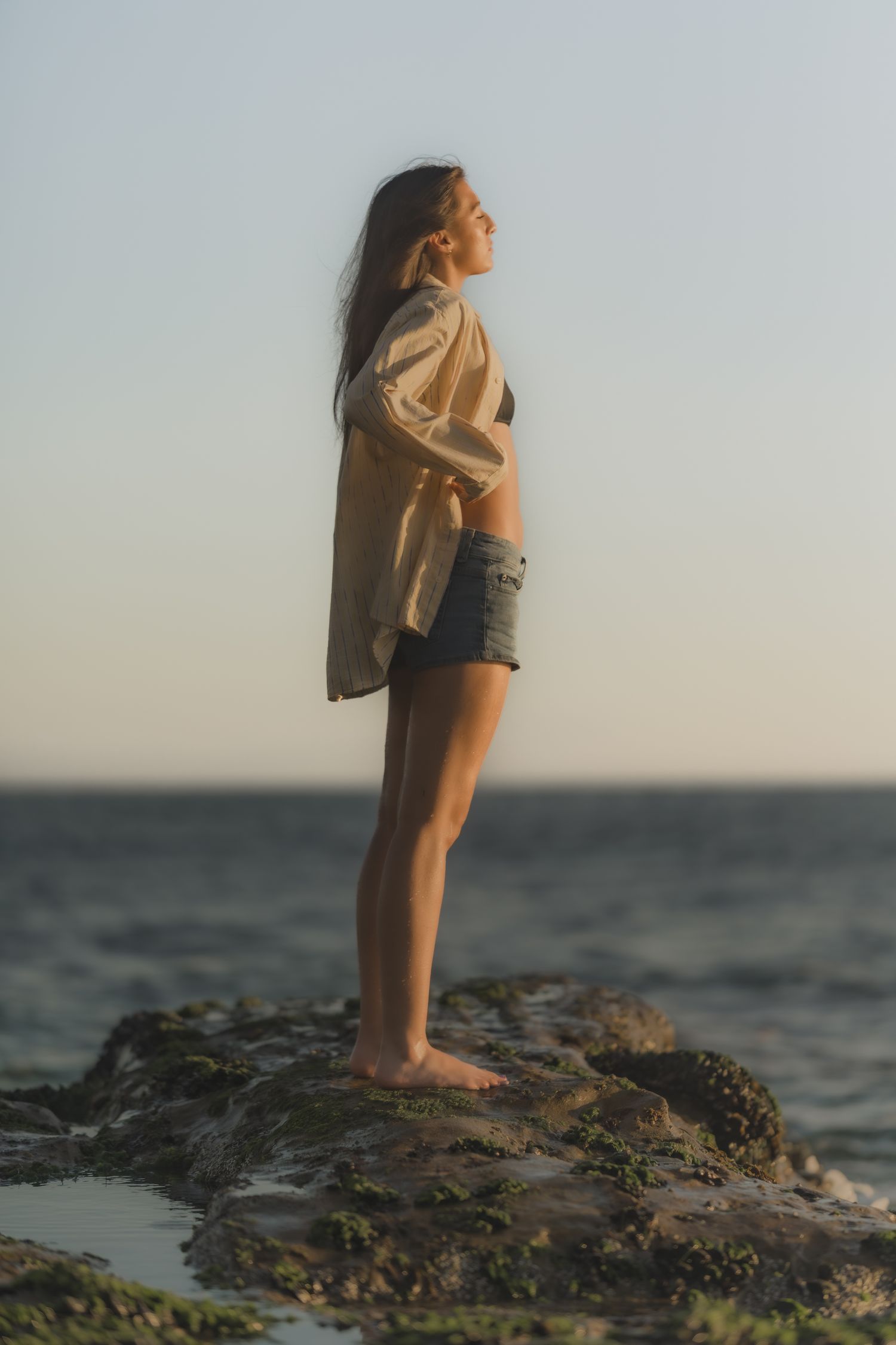 A barefoot woman in denim shorts and an open shirt stands on mossy rocks by the ocean at golden hour.